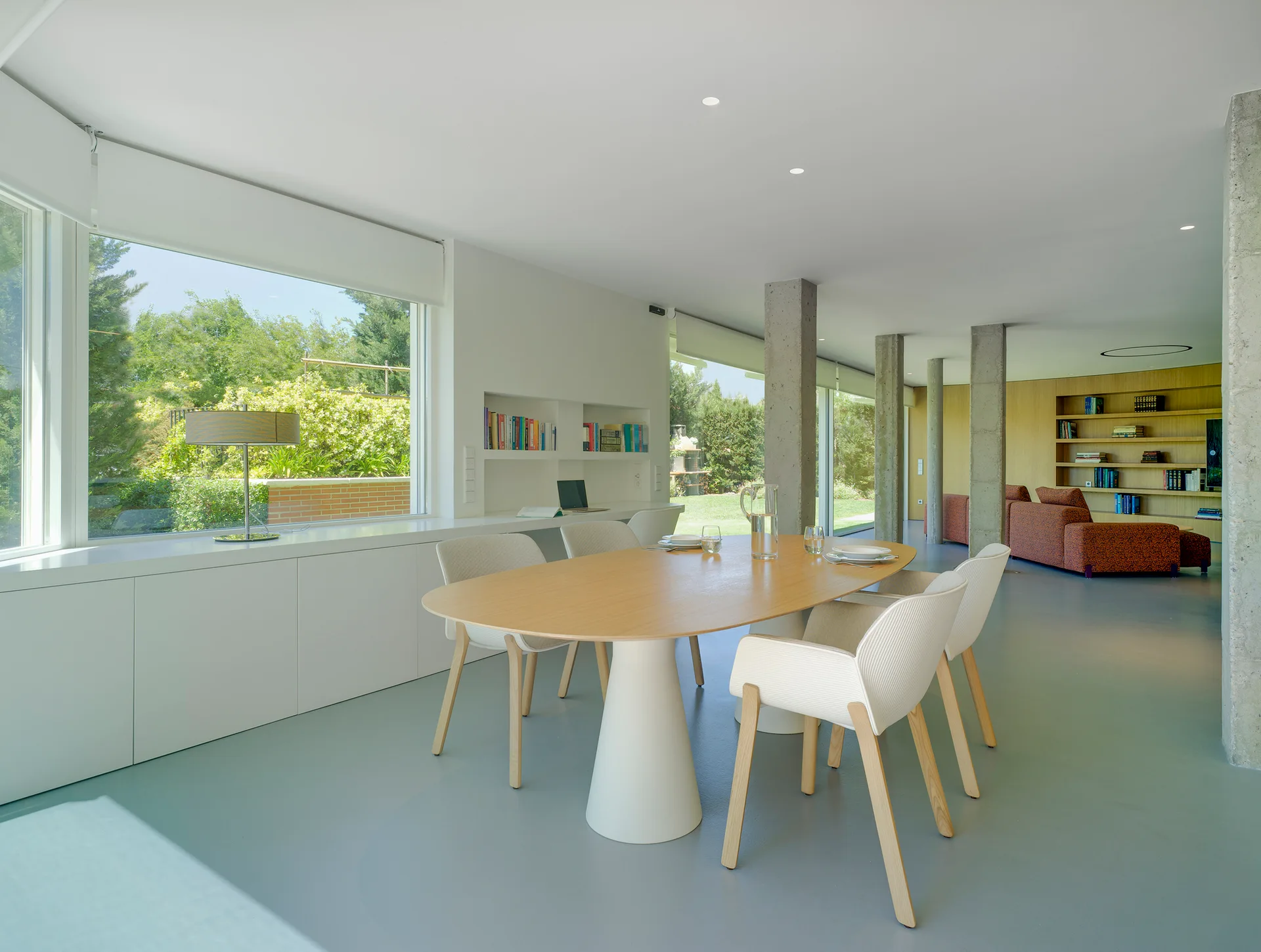 A minimalist open-plan dining room featuring an oval wood table, white chairs, and exposed concrete structural pillars.