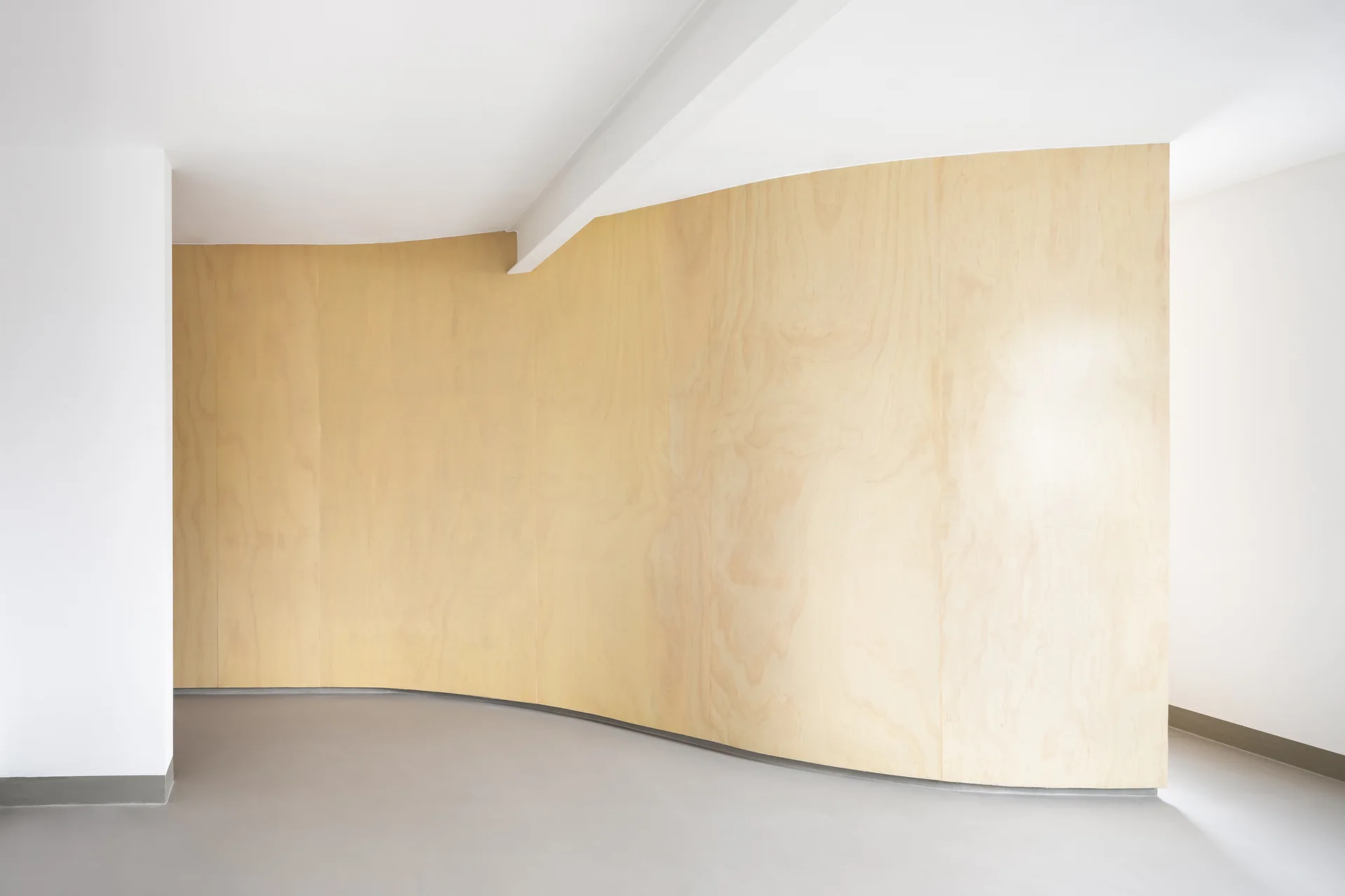 A minimalist room features a large curved partition wall covered in light wood plywood panels alongside white painted walls and a smooth grey floor.
