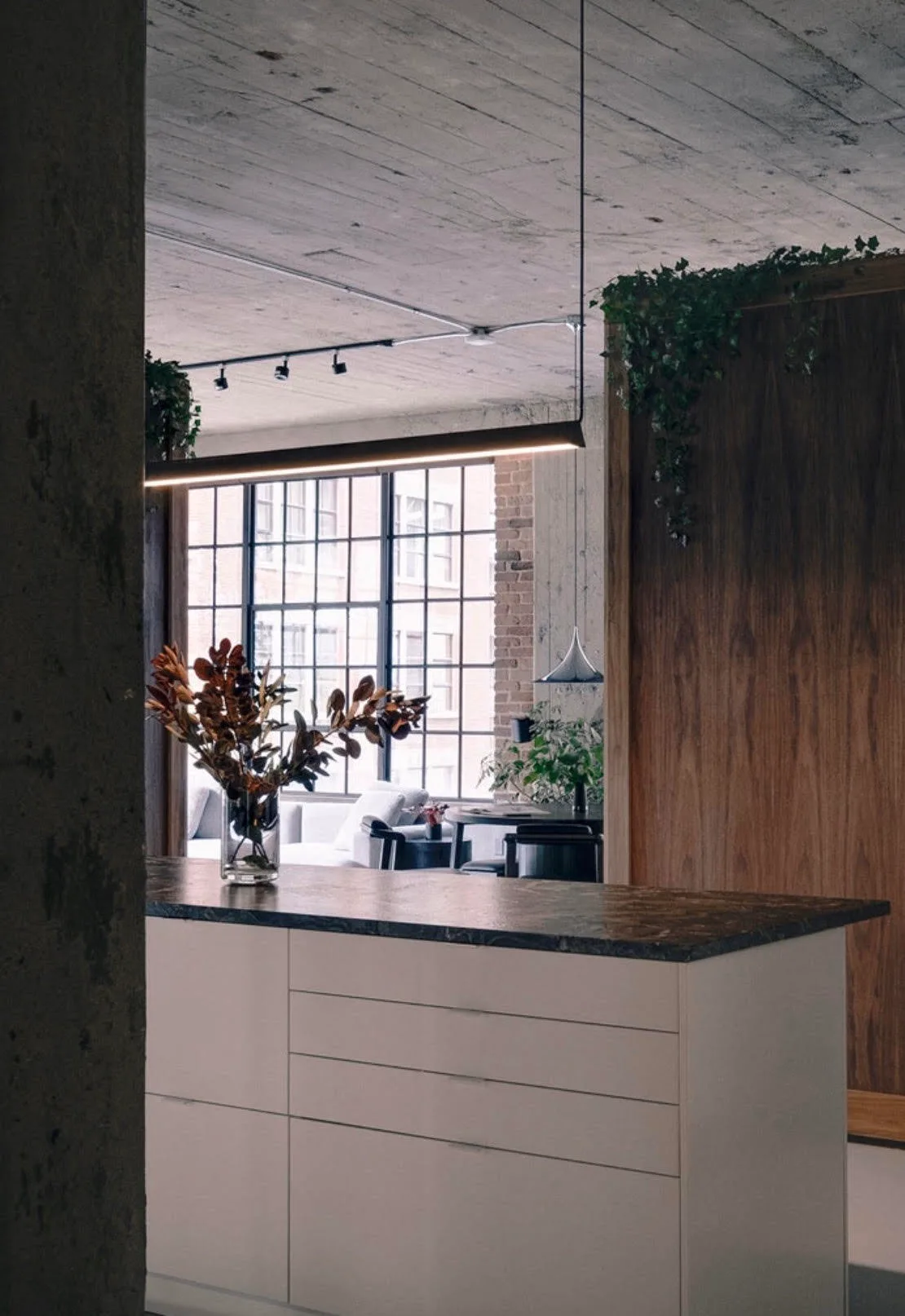 A modern kitchen island with a dark stone countertop sits below a linear pendant light, with a wood partition wall and grid window in the background.