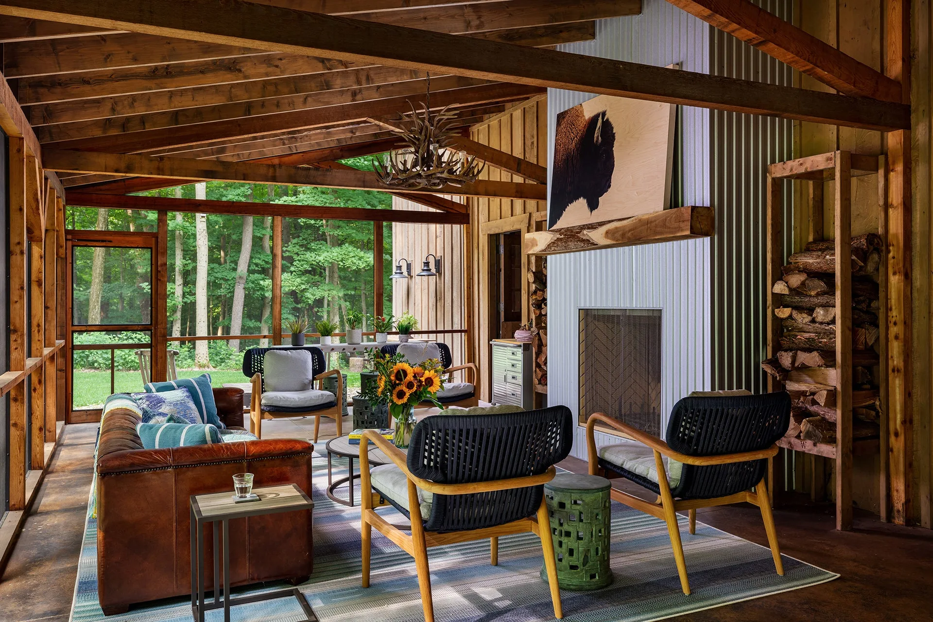 A rustic screened porch features a brown leather sofa, woven armchairs, and a corrugated metal fireplace under a timber-beamed ceiling.