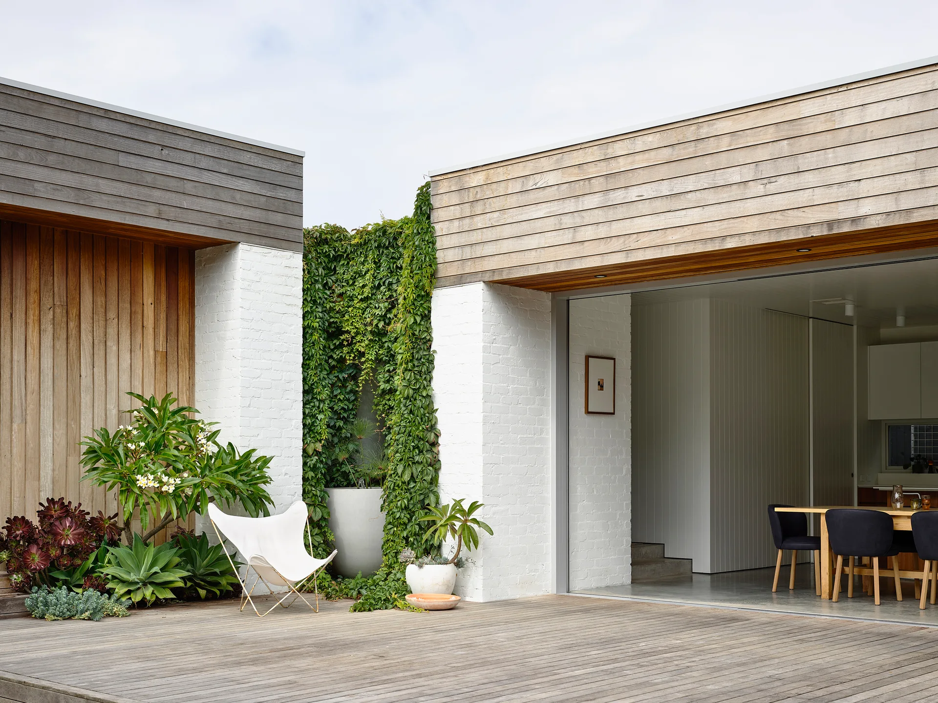 A modern house exterior featuring a wooden deck, white brick pillars, a butterfly chair, and an indoor dining room through open sliding doors.