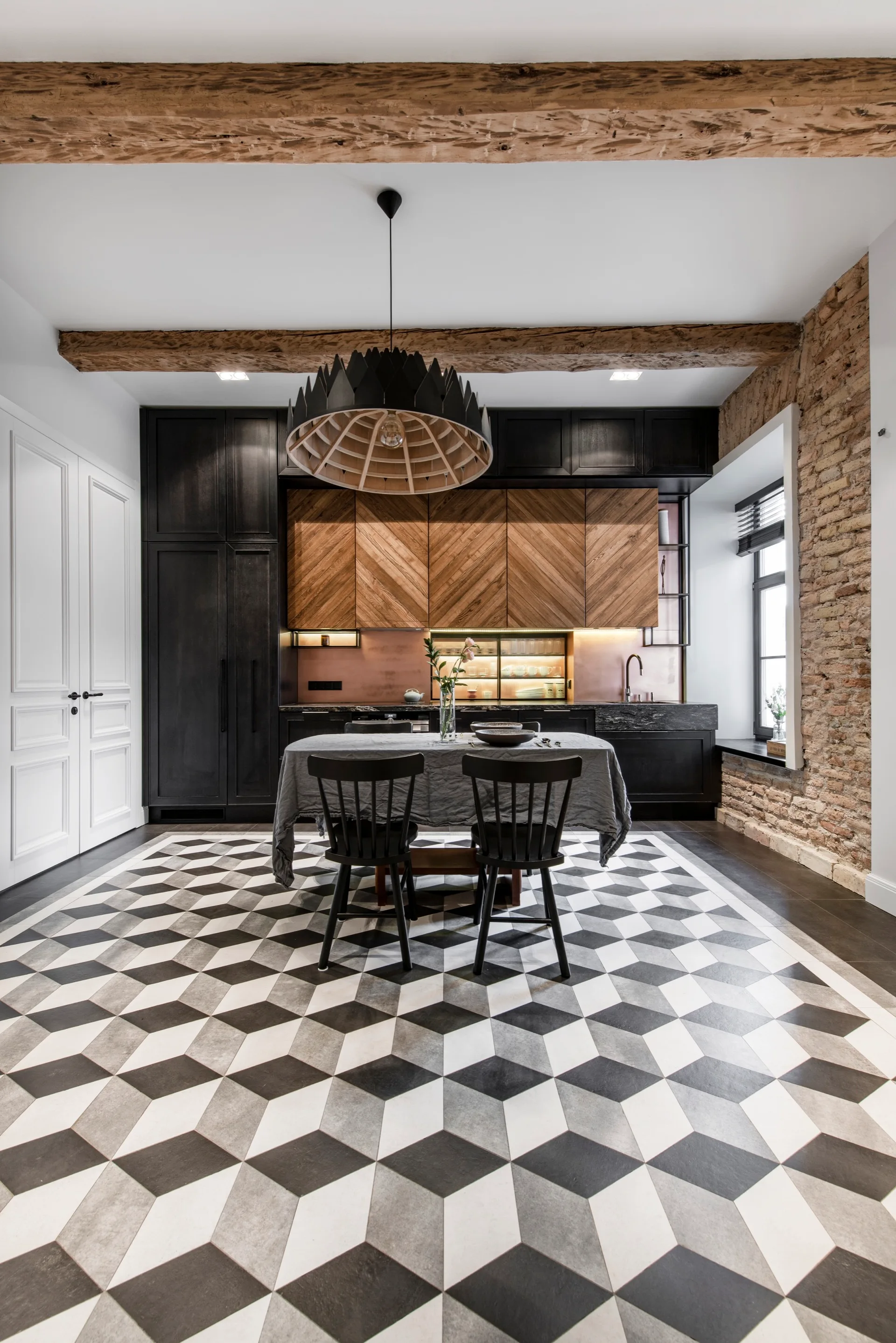 A modern kitchen features bold black-and-white geometric floor tiles, diagonal wood cabinet fronts, an exposed brick wall, and a central dining table.