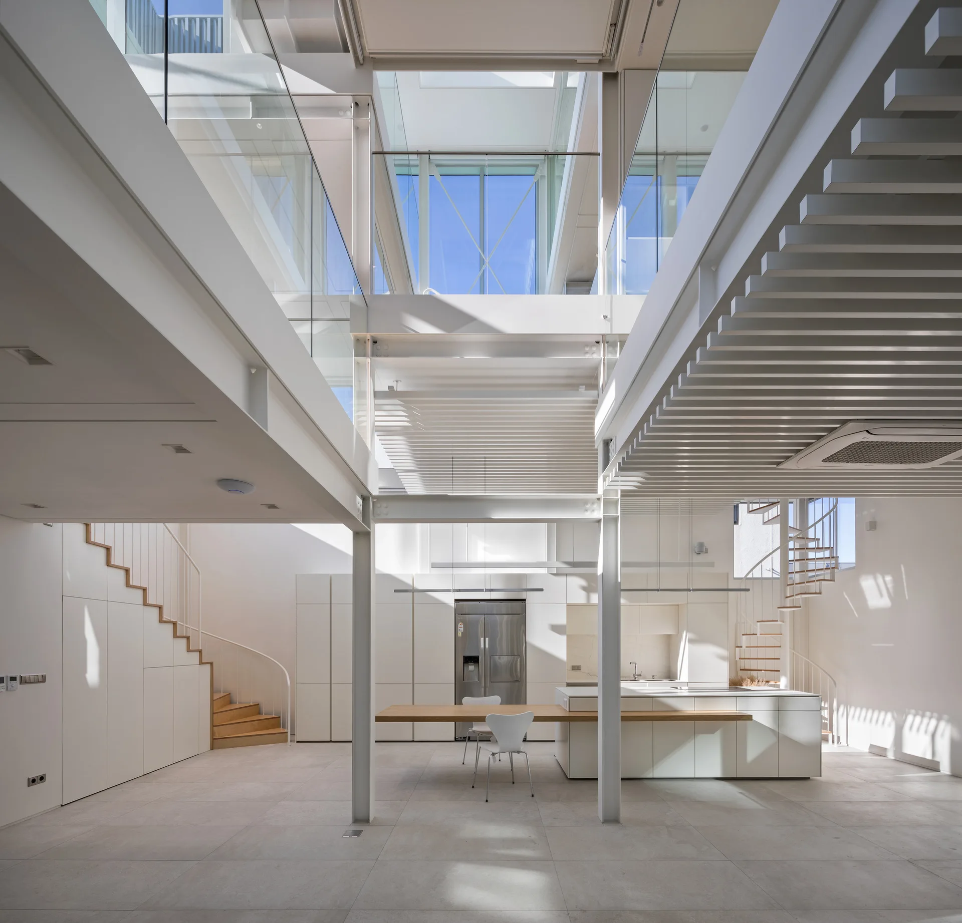 A double-height minimalist kitchen features a central white island with an attached wood table, built-in storage stairs, and overhead skylights.