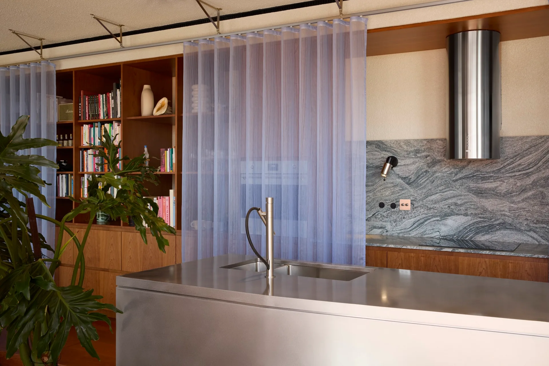 A contemporary kitchen featuring a stainless steel island, a textured stone backsplash, wooden shelves with books, and a sheer blue curtain.