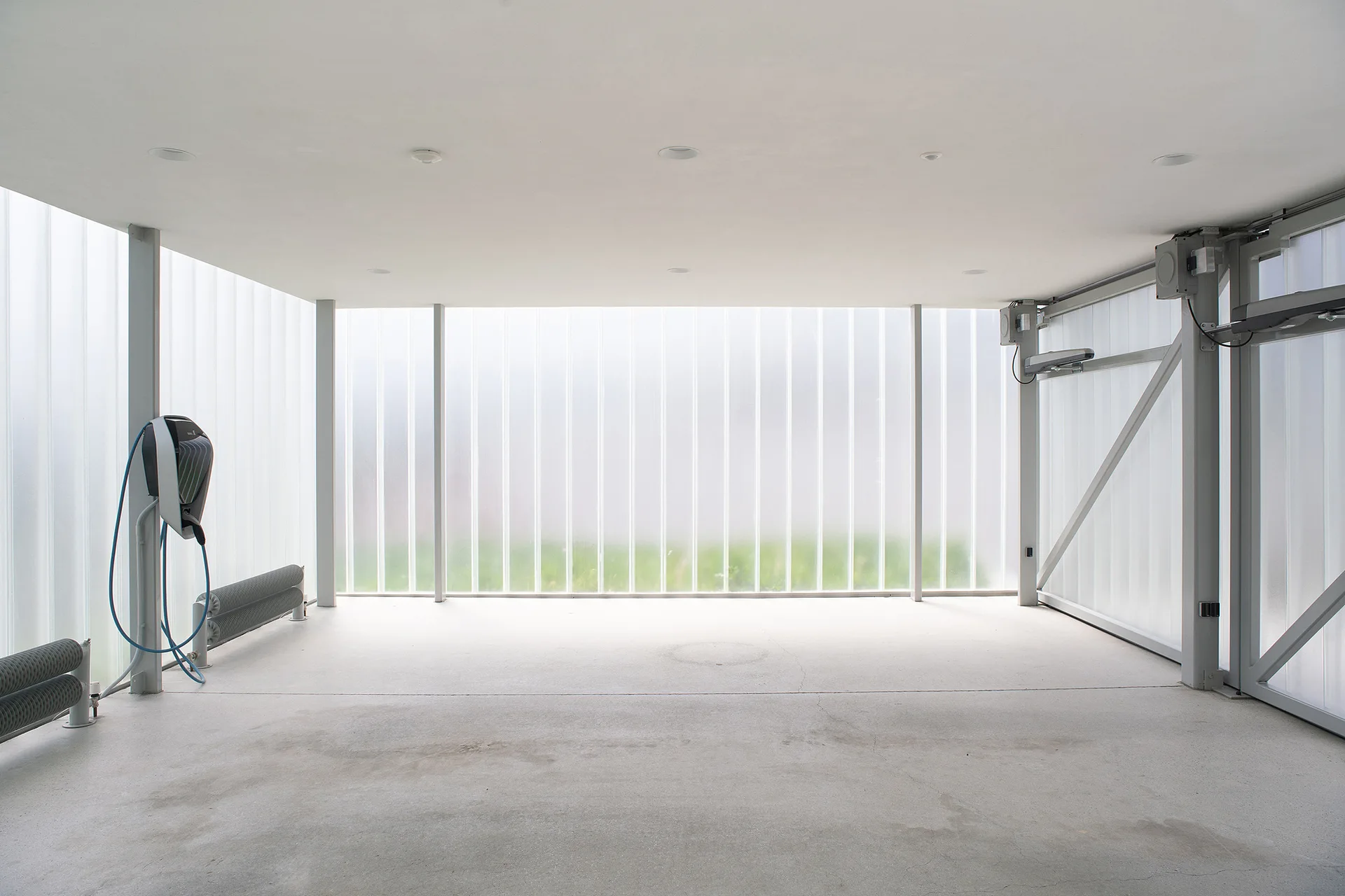 A minimalist utility garage featuring translucent ribbed glass walls, concrete floors, and a wall-mounted electric vehicle charging station.