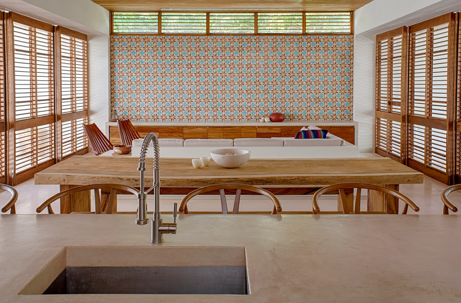 An open-concept kitchen and living area features a concrete island in the foreground, a wooden dining table, and a geometric tiled accent wall.