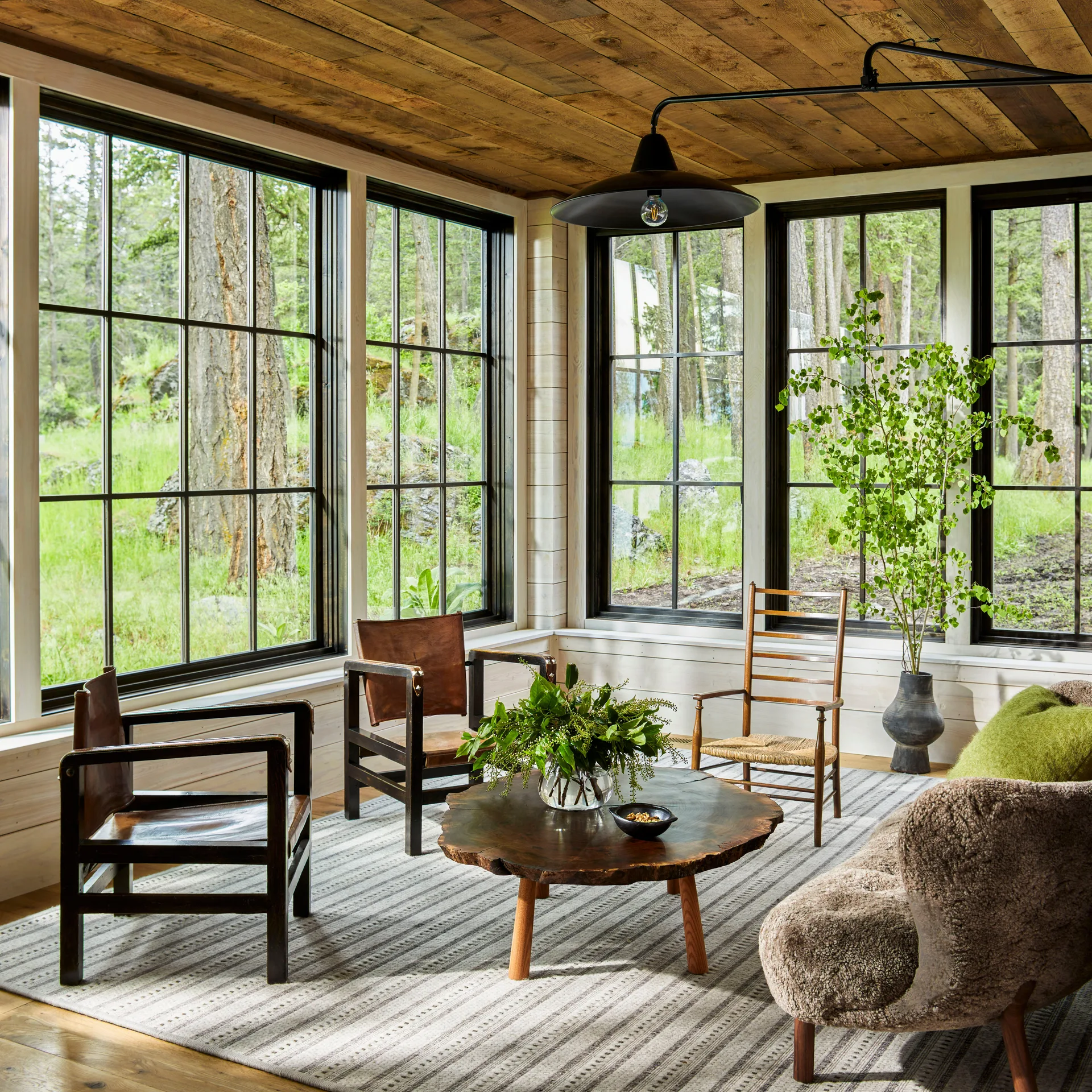 A living room seating area features a brown sheepskin sofa, leather armchairs, a live-edge wooden coffee table, and large black-framed windows.
