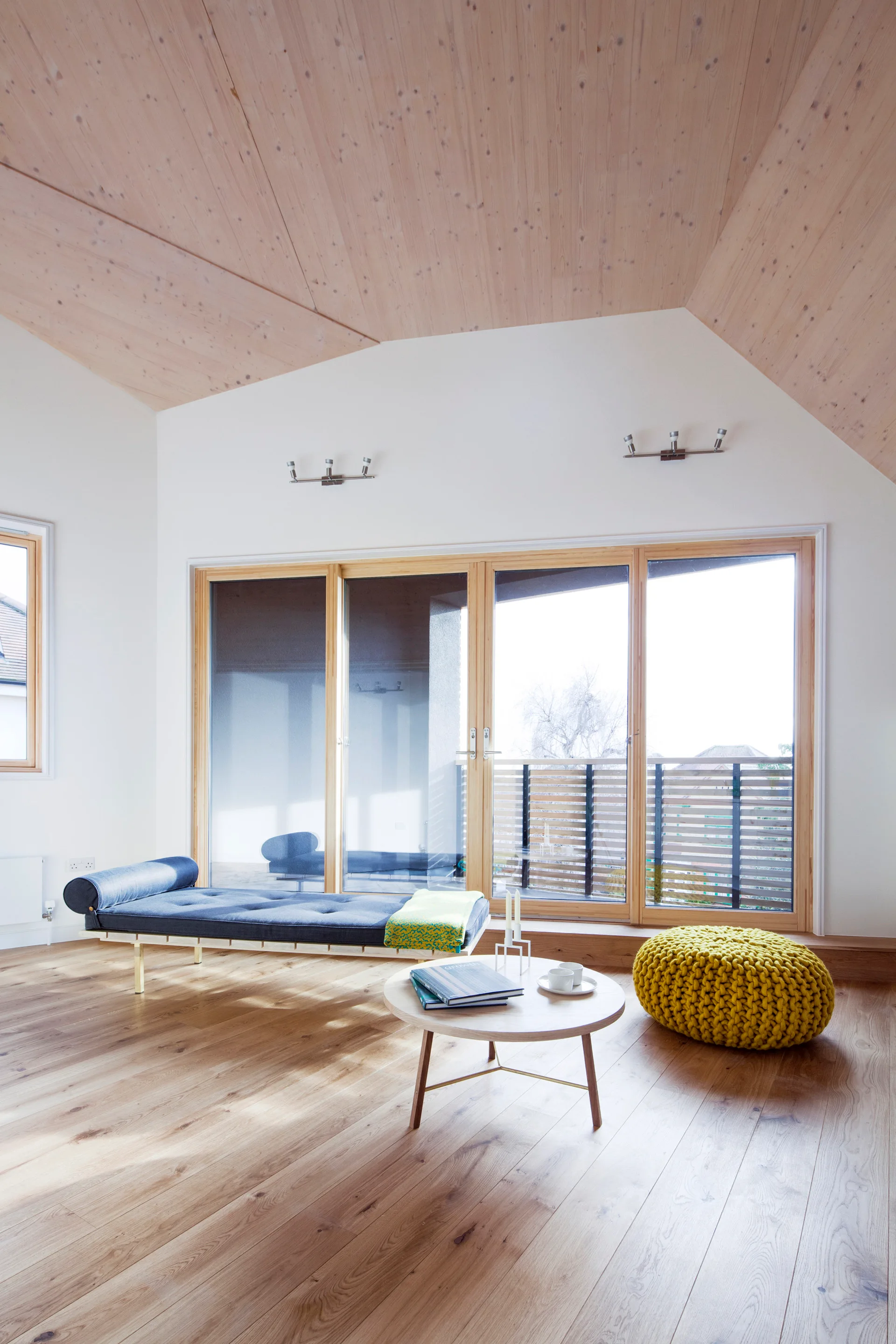 A minimalist living room with a pitched wood-paneled ceiling, a blue upholstered daybed, a round coffee table, and a yellow knit pouf.