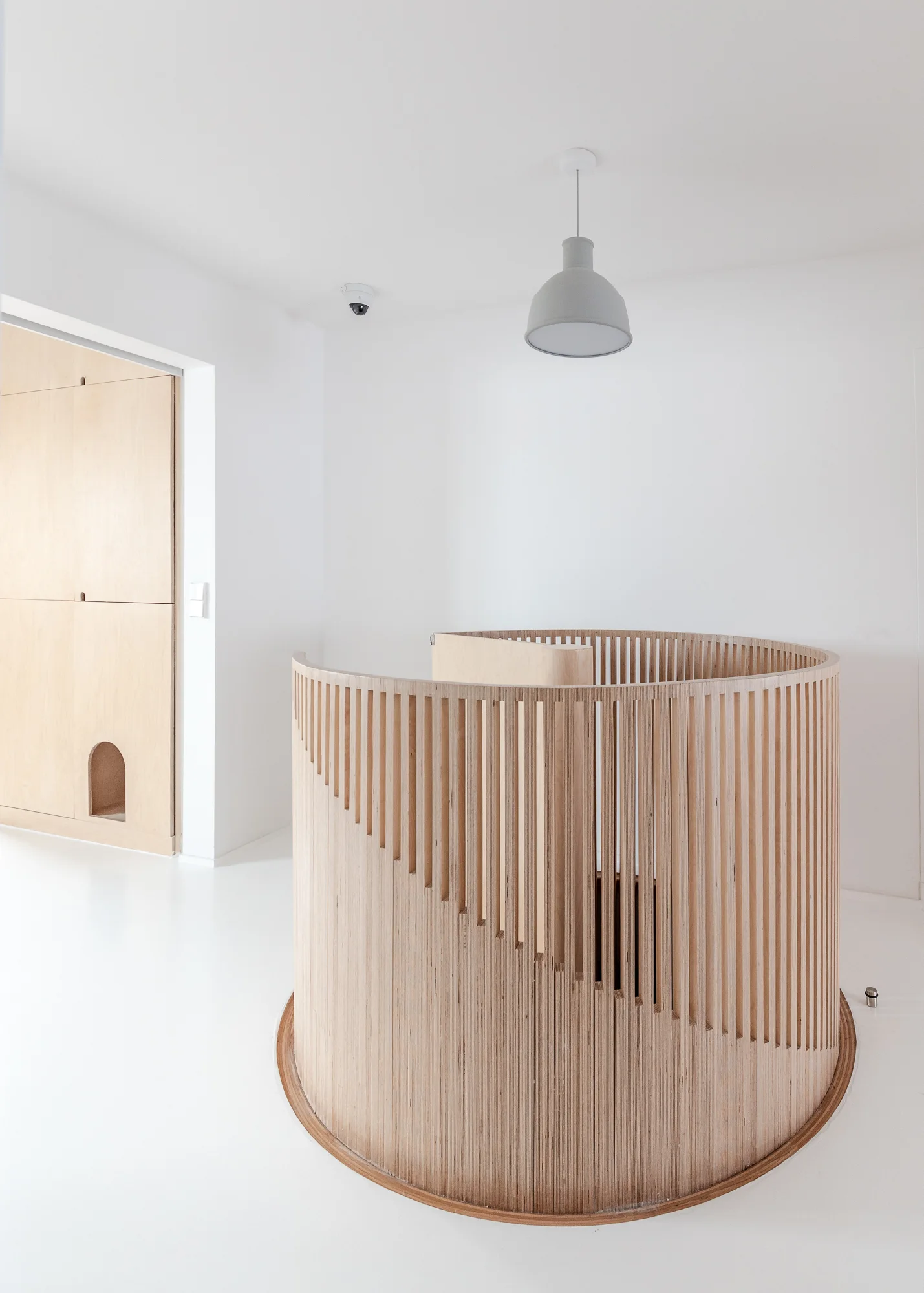 A circular slatted light wood staircase railing stands on a seamless white floor next to matching plywood wall paneling and a gray pendant light.