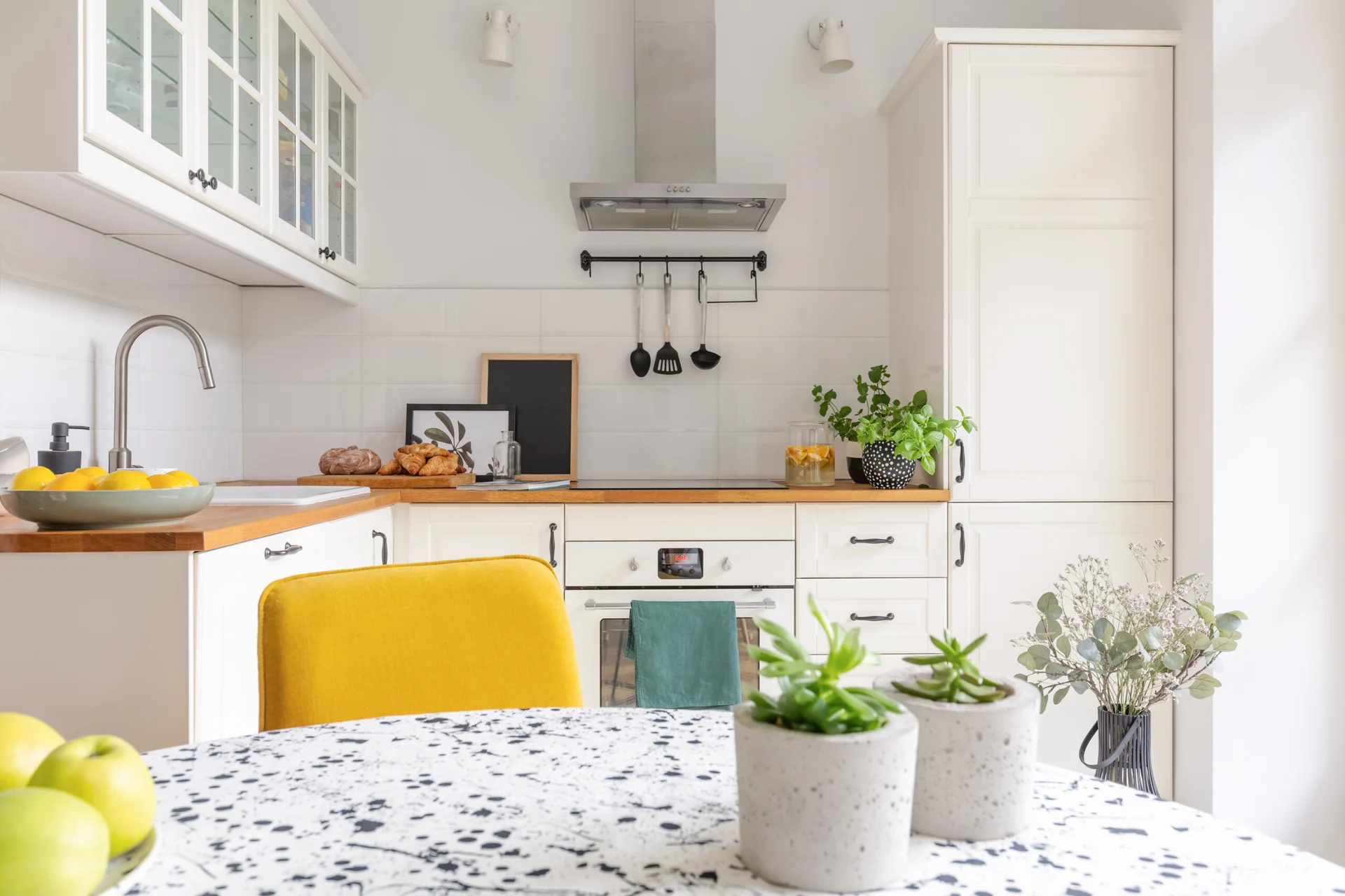 A bright kitchen features white cabinetry with wood countertops, a stainless steel range hood, and a dining table with a yellow chair in the foreground.
