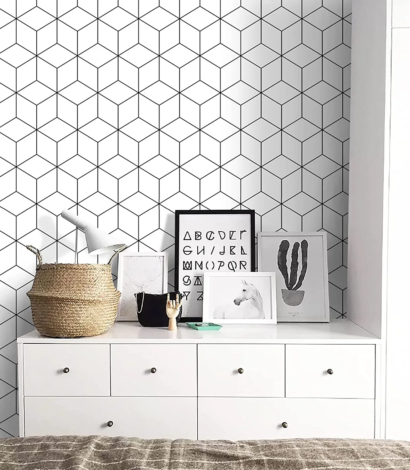 A white dresser featuring layered framed art prints and a woven basket is set against black and white geometric cube wallpaper.