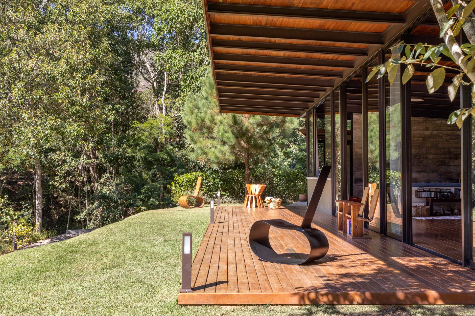 A modern wooden deck with sculptural chairs is flanked by floor-to-ceiling glass doors and a lush green forest backdrop under exposed rafters.