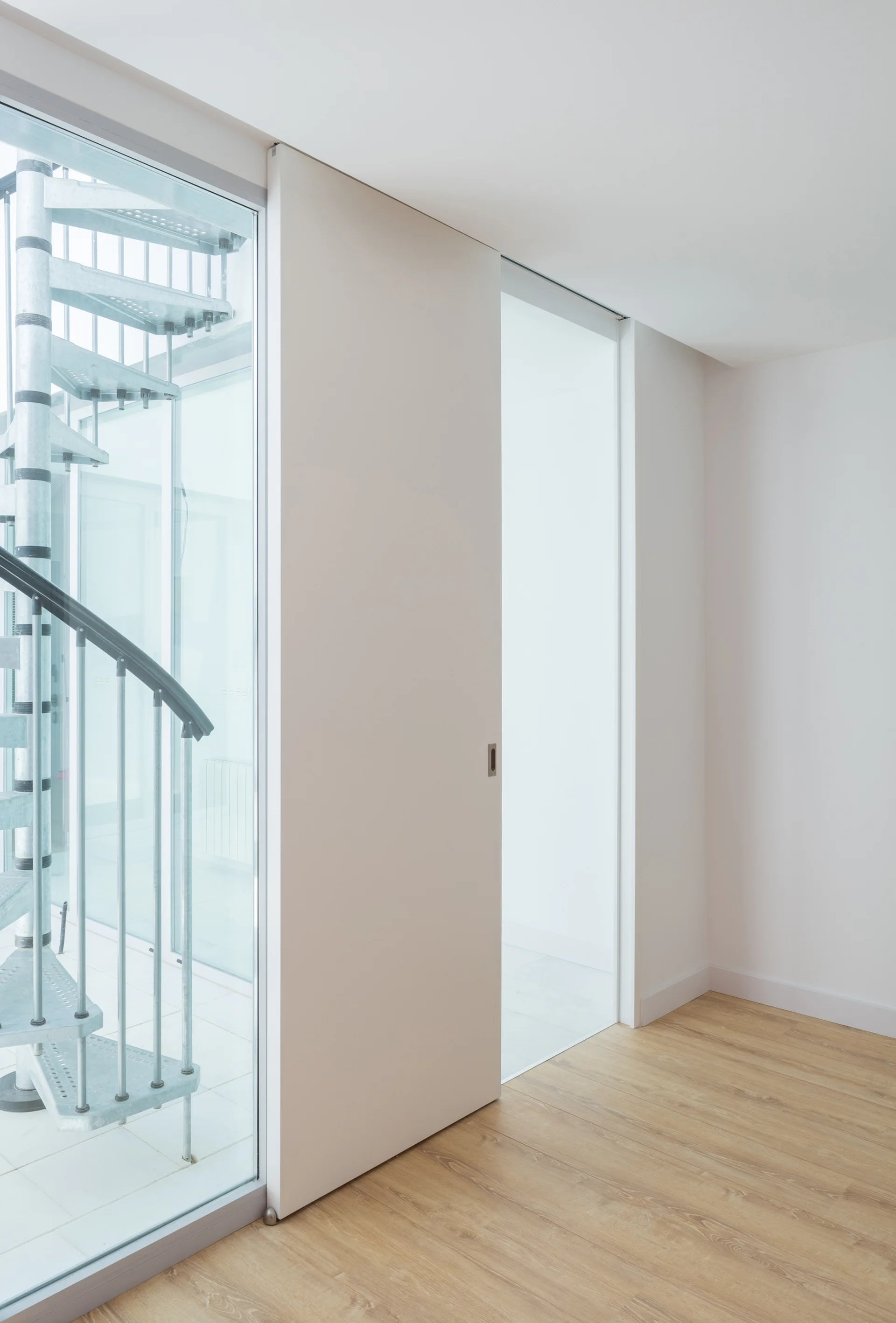 A minimalist white room features light wood floors, a large white sliding door, and a metal spiral staircase visible behind a glass partition.