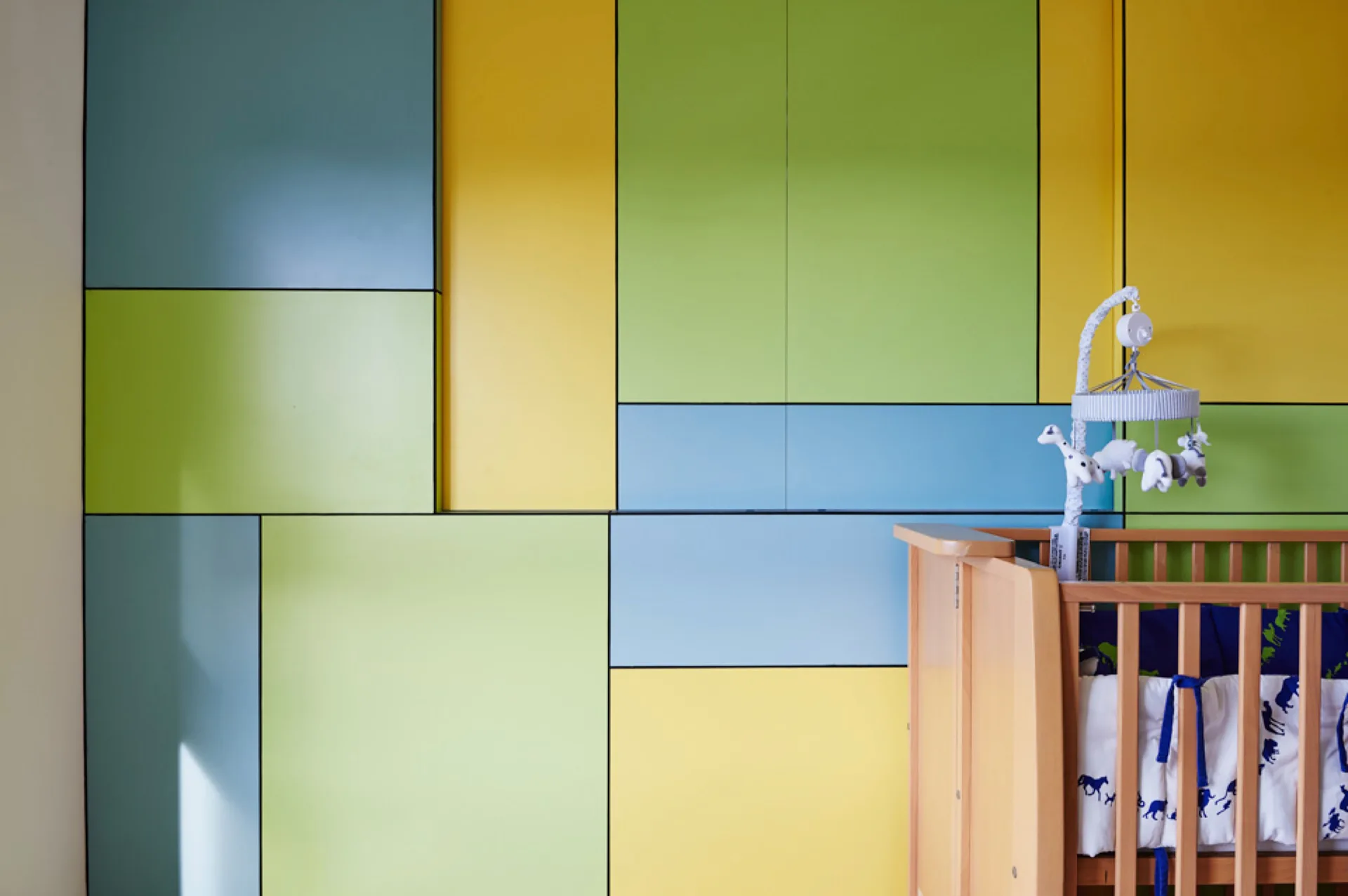 A wooden baby crib with a mobile stands in front of a flat-panel wall decorated in large blue, green, and yellow geometric blocks.
