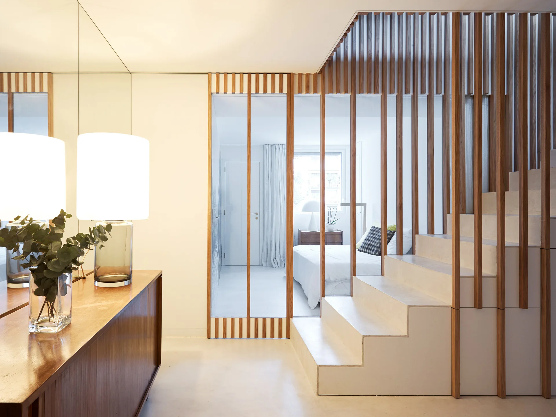 A modern white staircase features a vertical wood slat balustrade that integrates into a glass room divider separating the hallway from a bedroom.