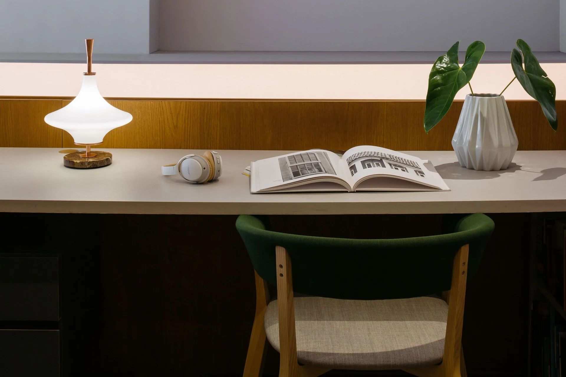 A modern grey desk features a glowing mid-century lamp, open book, white headphones, a geometric vase with green leaves, and a green upholstered chair.