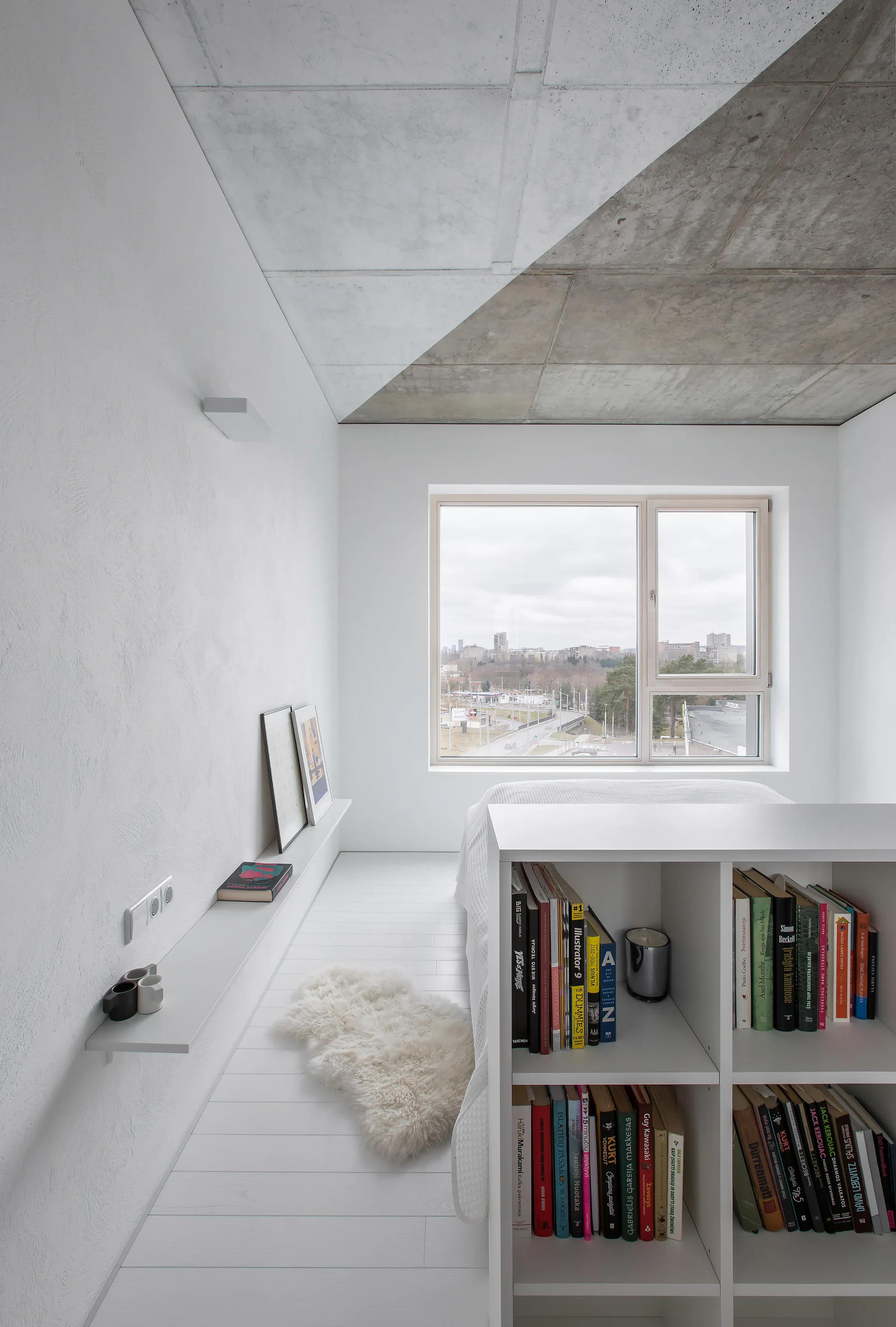 A minimalist white bedroom features an exposed concrete ceiling, a low white wall shelf, and a bookshelf room divider next to a large window.