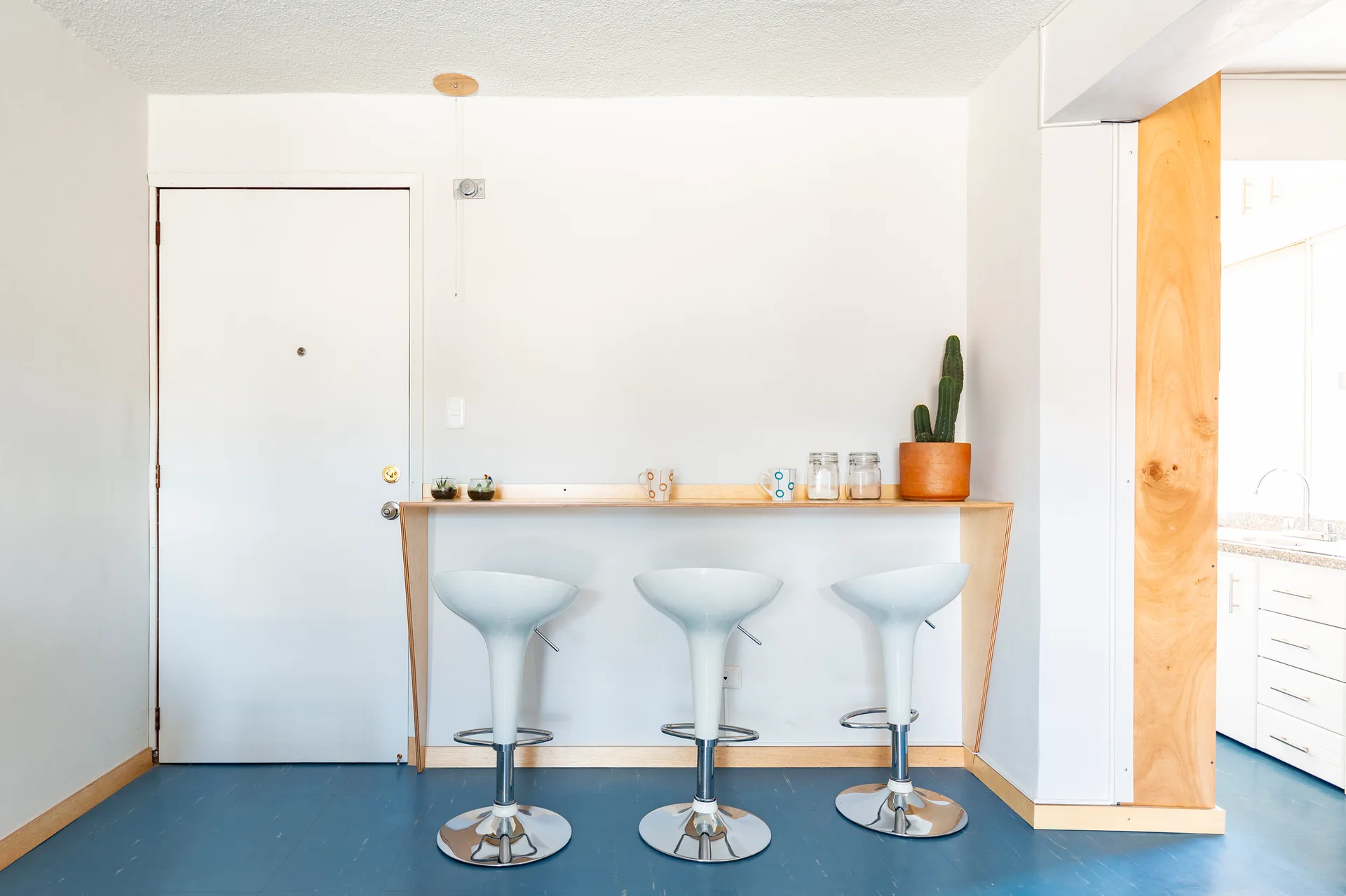 Three white pedestal bar stools sit at a slim wooden breakfast bar mounted on a wall above a solid blue floor.