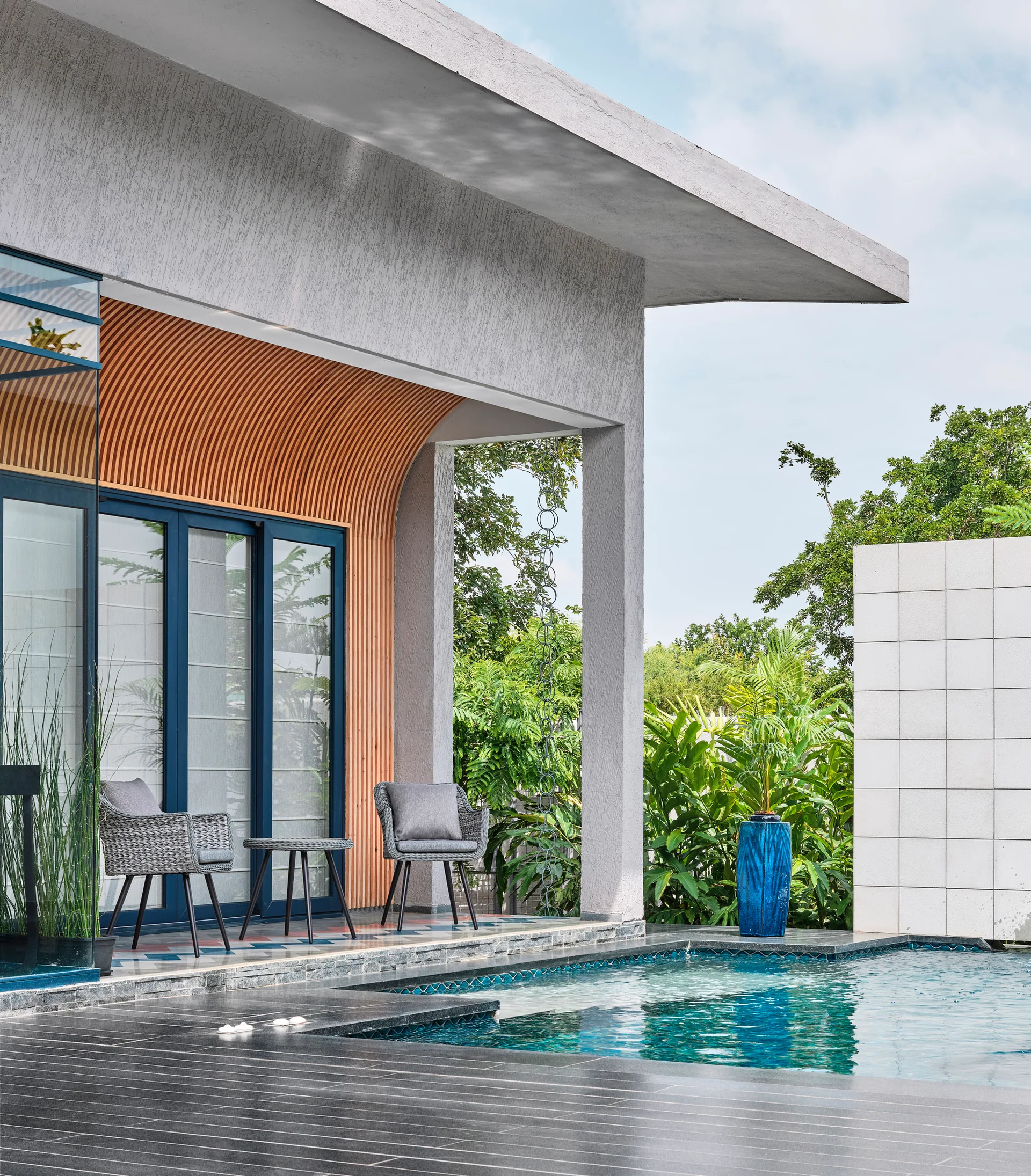 A modern outdoor pool deck features gray woven patio chairs beside a swimming pool, set against a curved wood-slat wall and a textured concrete overhang.