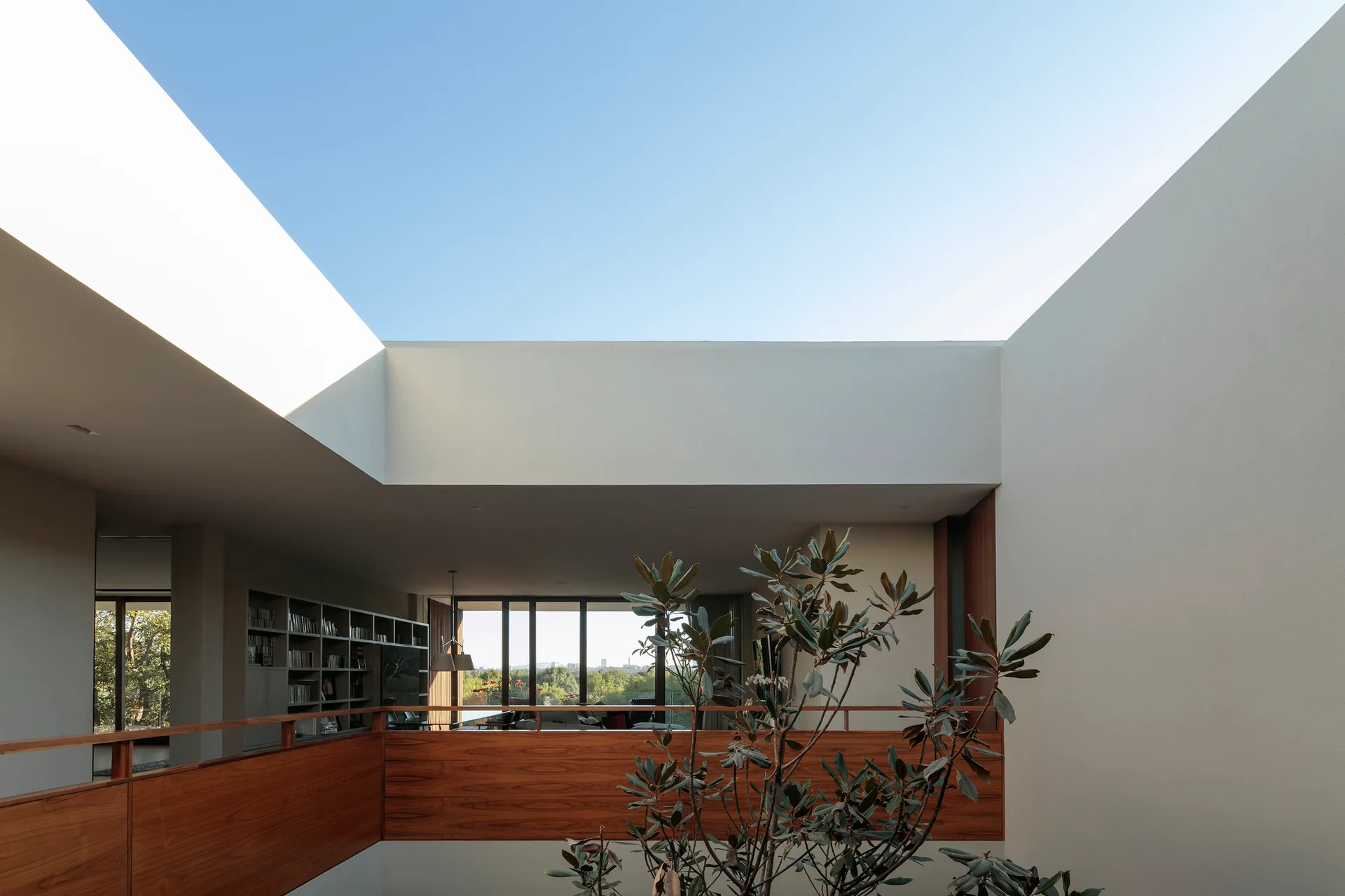 An open-air atrium features a warm wood mezzanine railing overlooking an interior living area with built-in bookshelves.