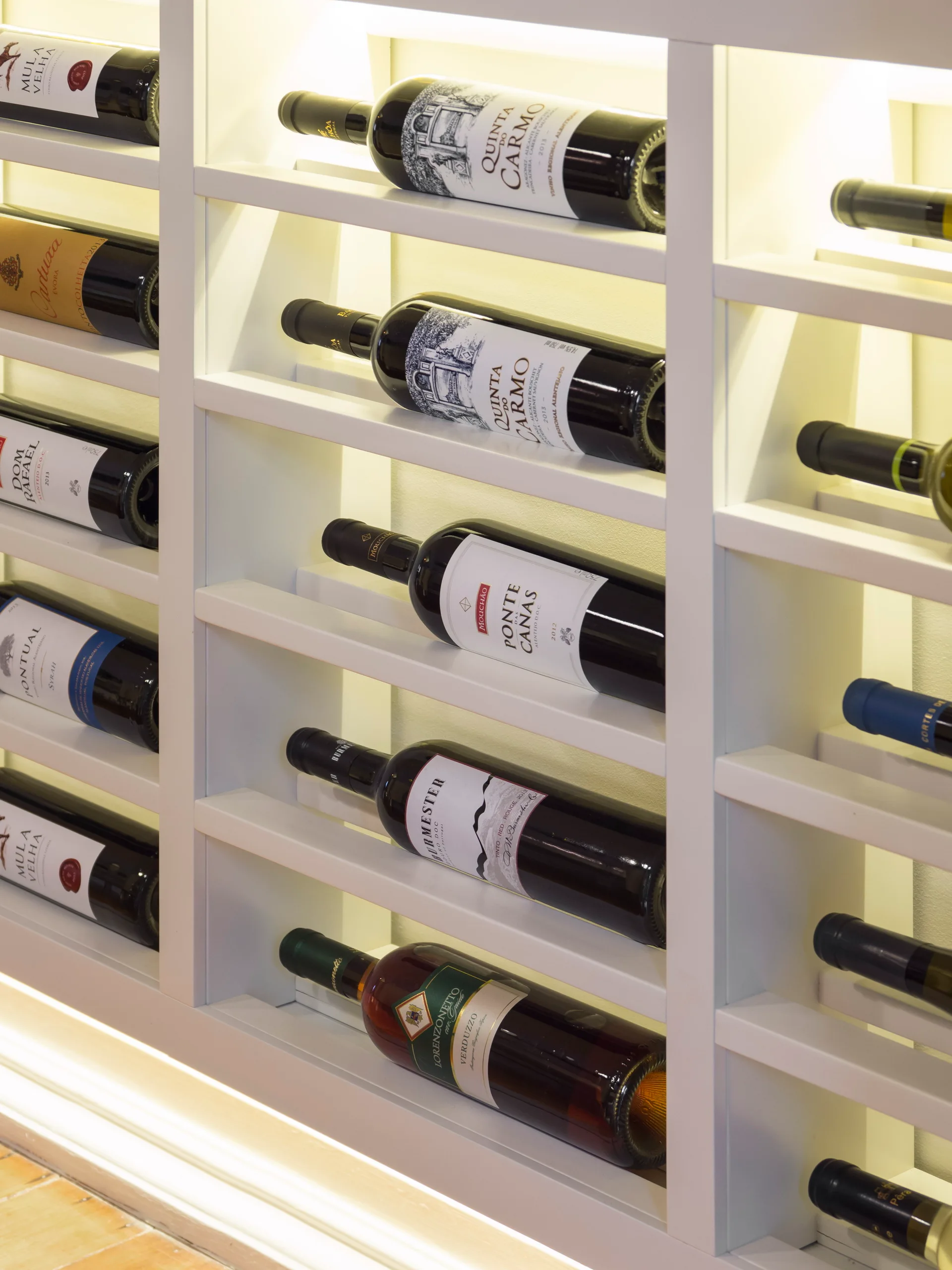 A close-up of a custom white wooden wine rack with multiple glass bottles stored horizontally on illuminated shelves.