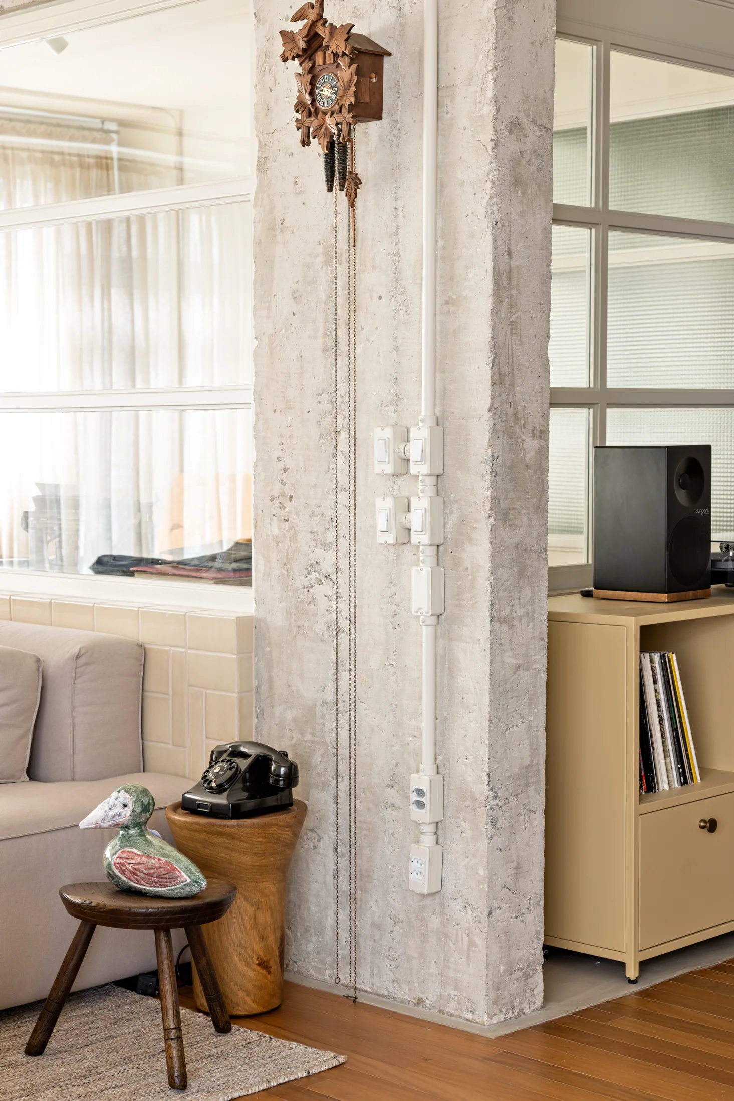 An eclectic living room features a raw concrete column holding a cuckoo clock, situated between a beige sofa and a metal record cabinet.