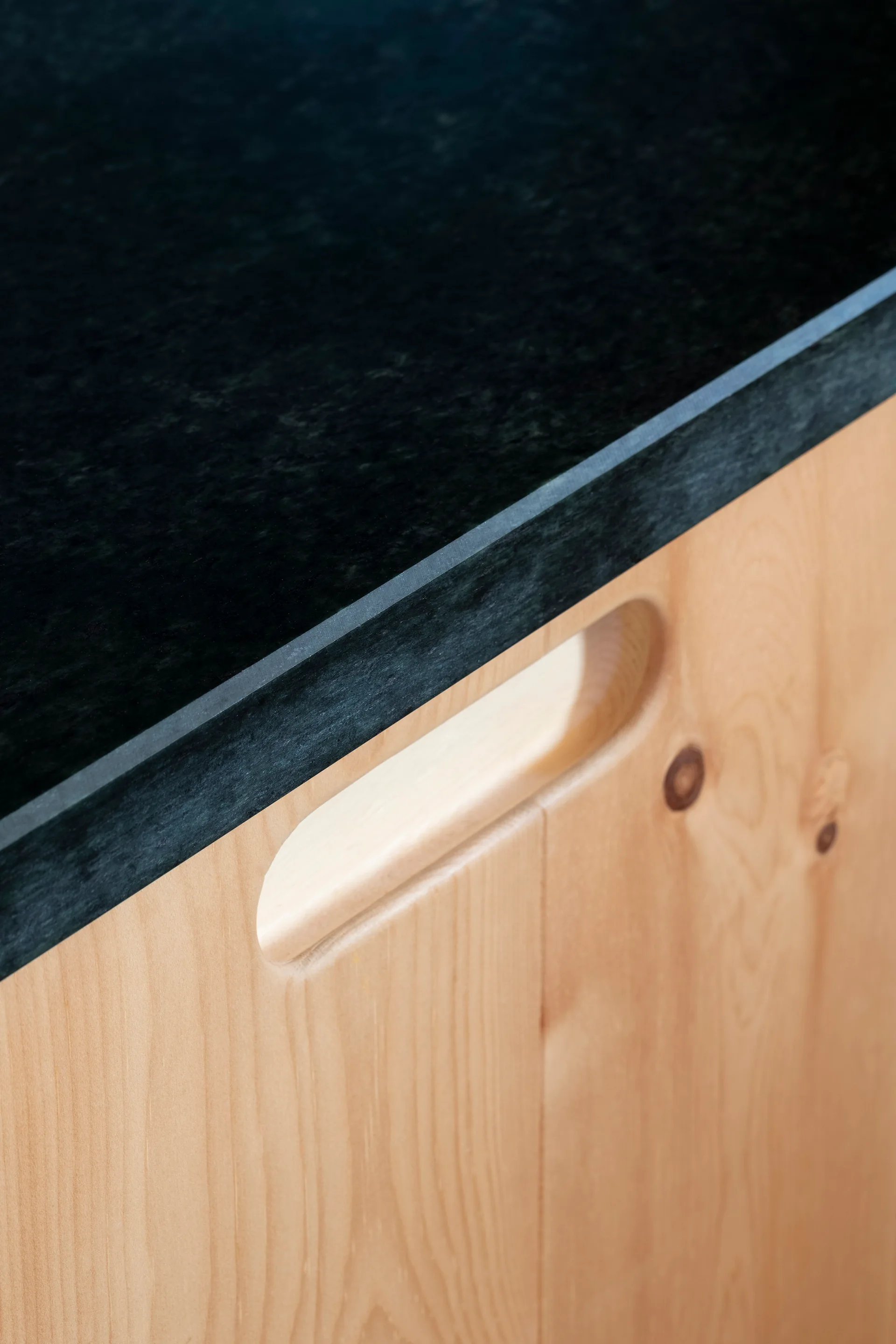 Close-up of a light wood kitchen cabinet featuring an integrated oval handle carved into the door under a dark stone countertop.