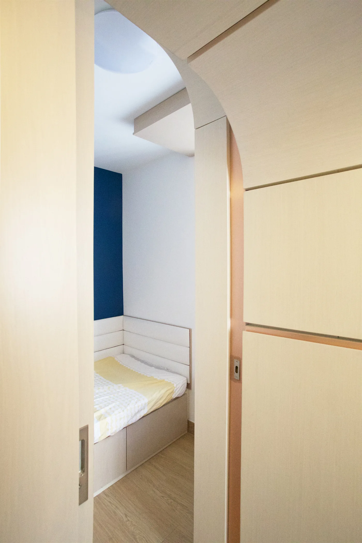 A view through a light wood arched doorway into a small bedroom with a corner bed, blue accent wall, and patterned bedding.