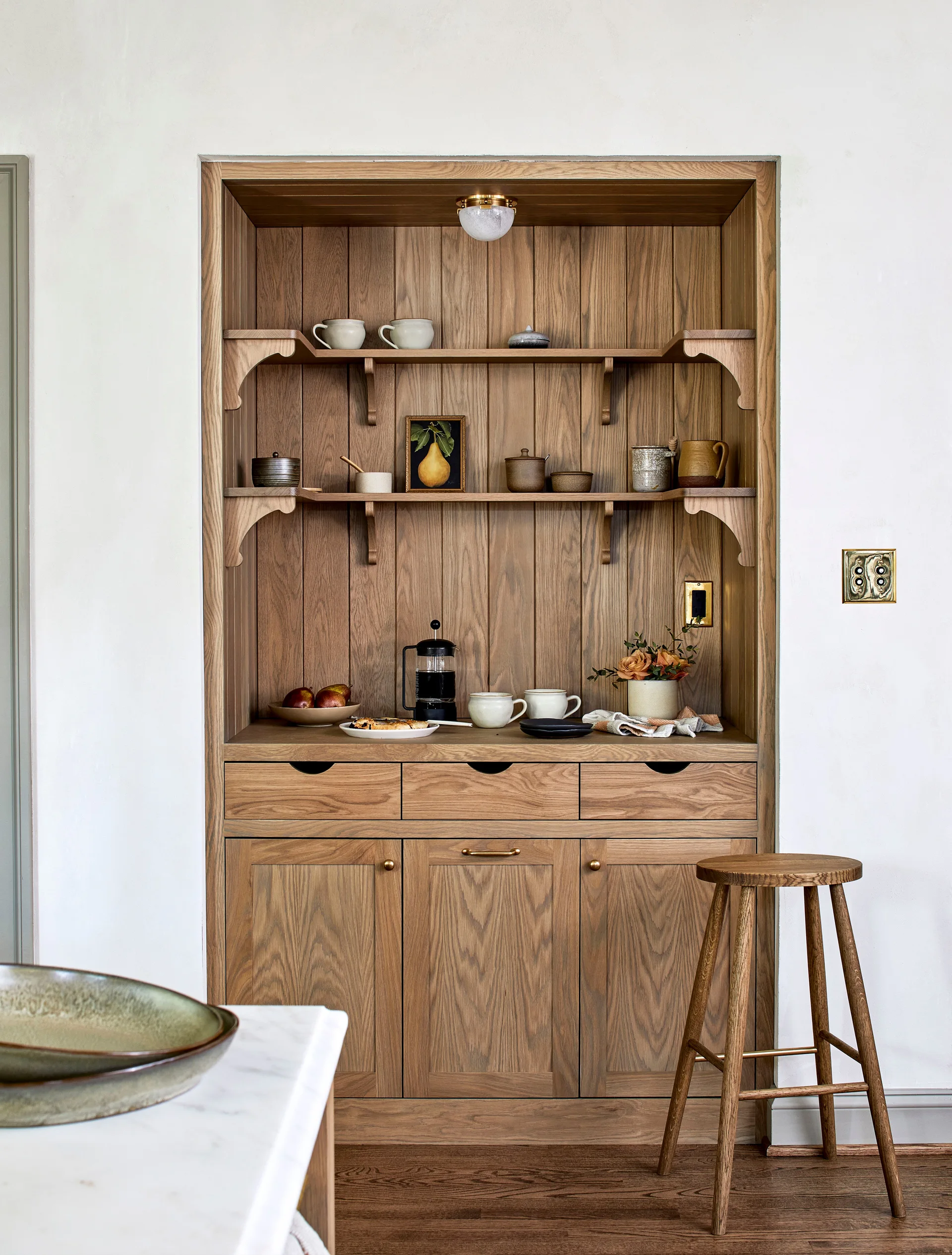 A recessed oak wood pantry cabinet built into a white plaster wall, featuring open shelving, brass accents, and a matching wooden stool.