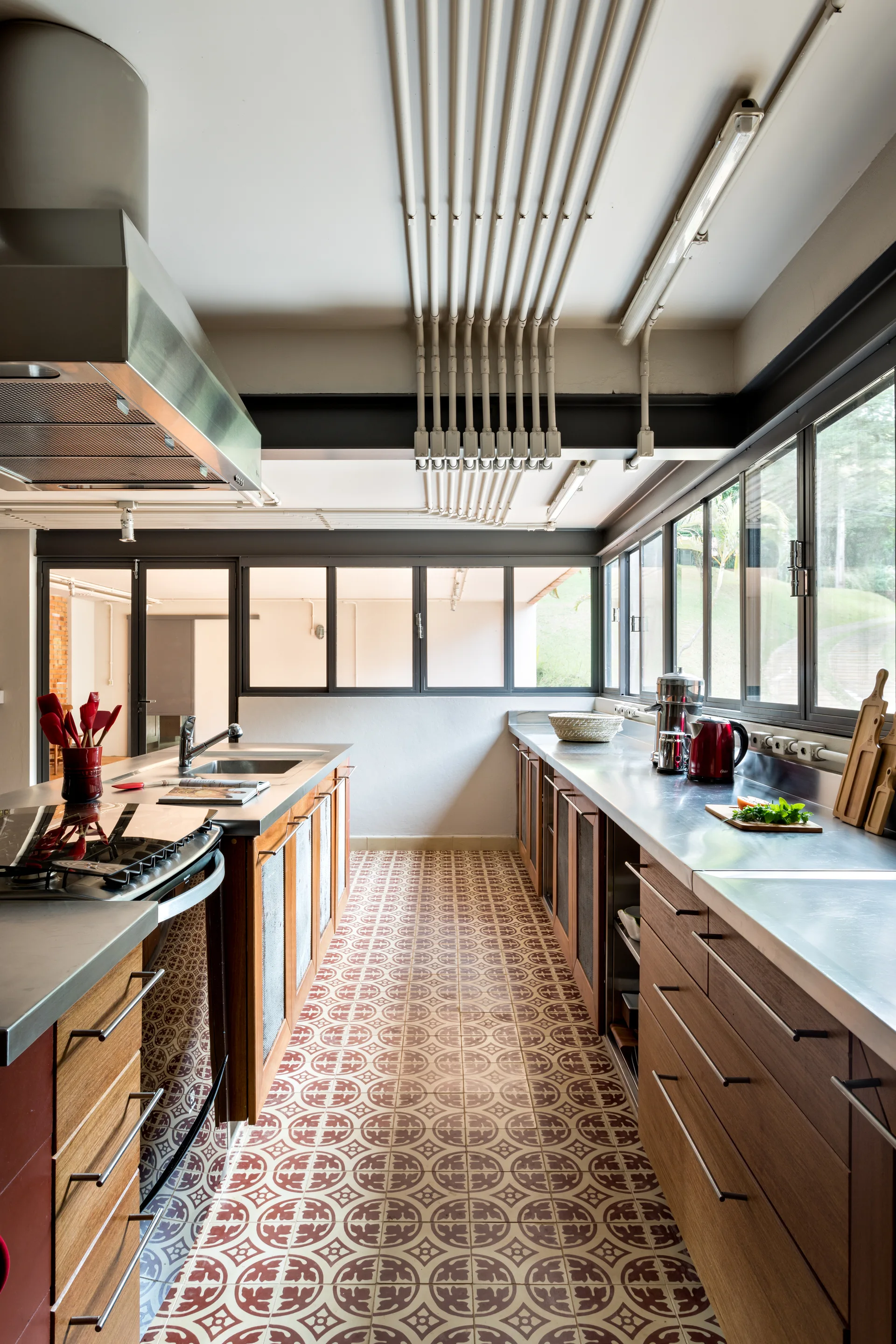 An industrial galley kitchen featuring stainless steel countertops, wood cabinets, patterned tile flooring, and exposed ceiling conduits.