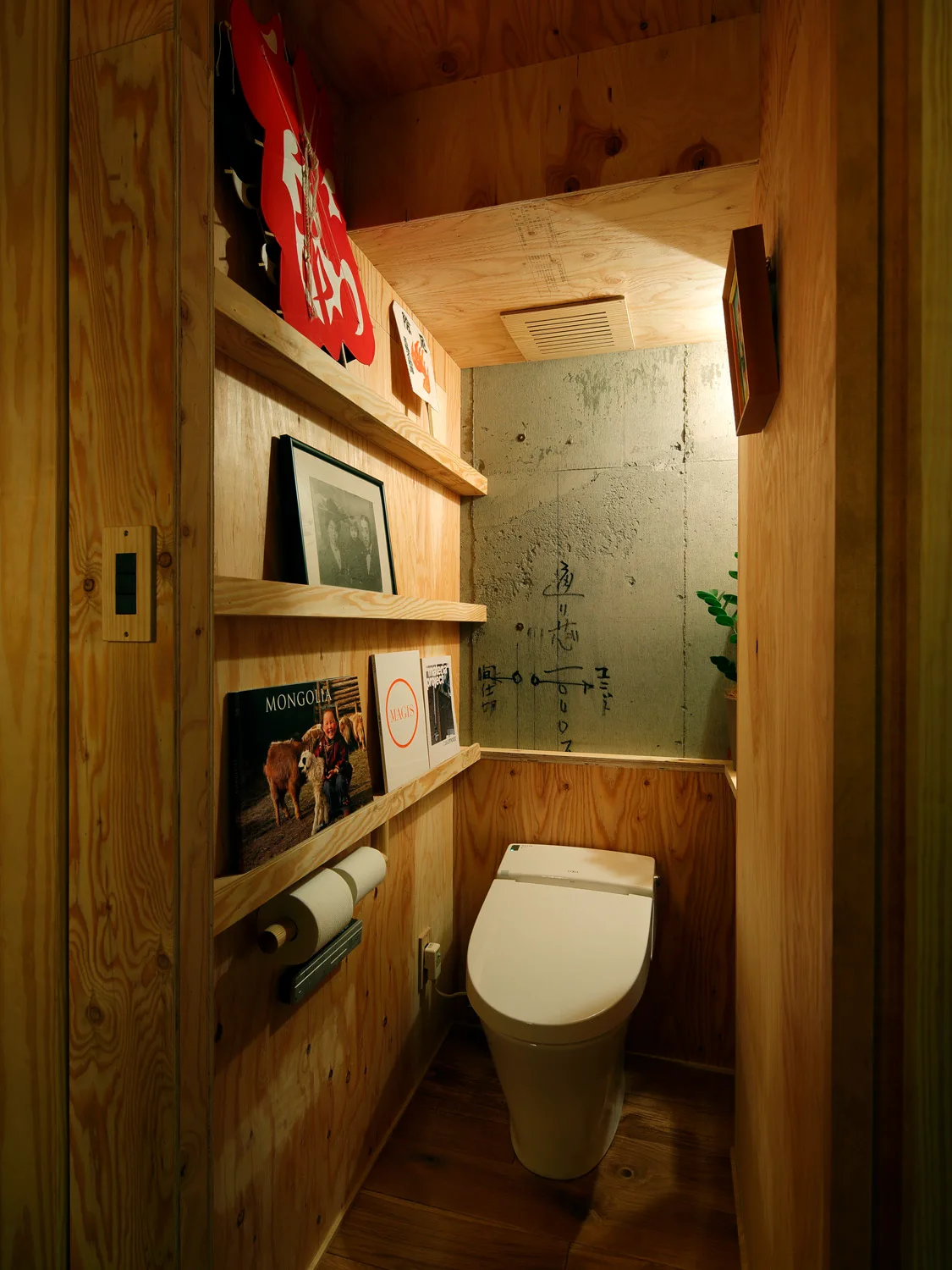 A compact powder room features a white toilet set against a raw concrete accent wall and surrounding plywood paneling with wooden display shelves.