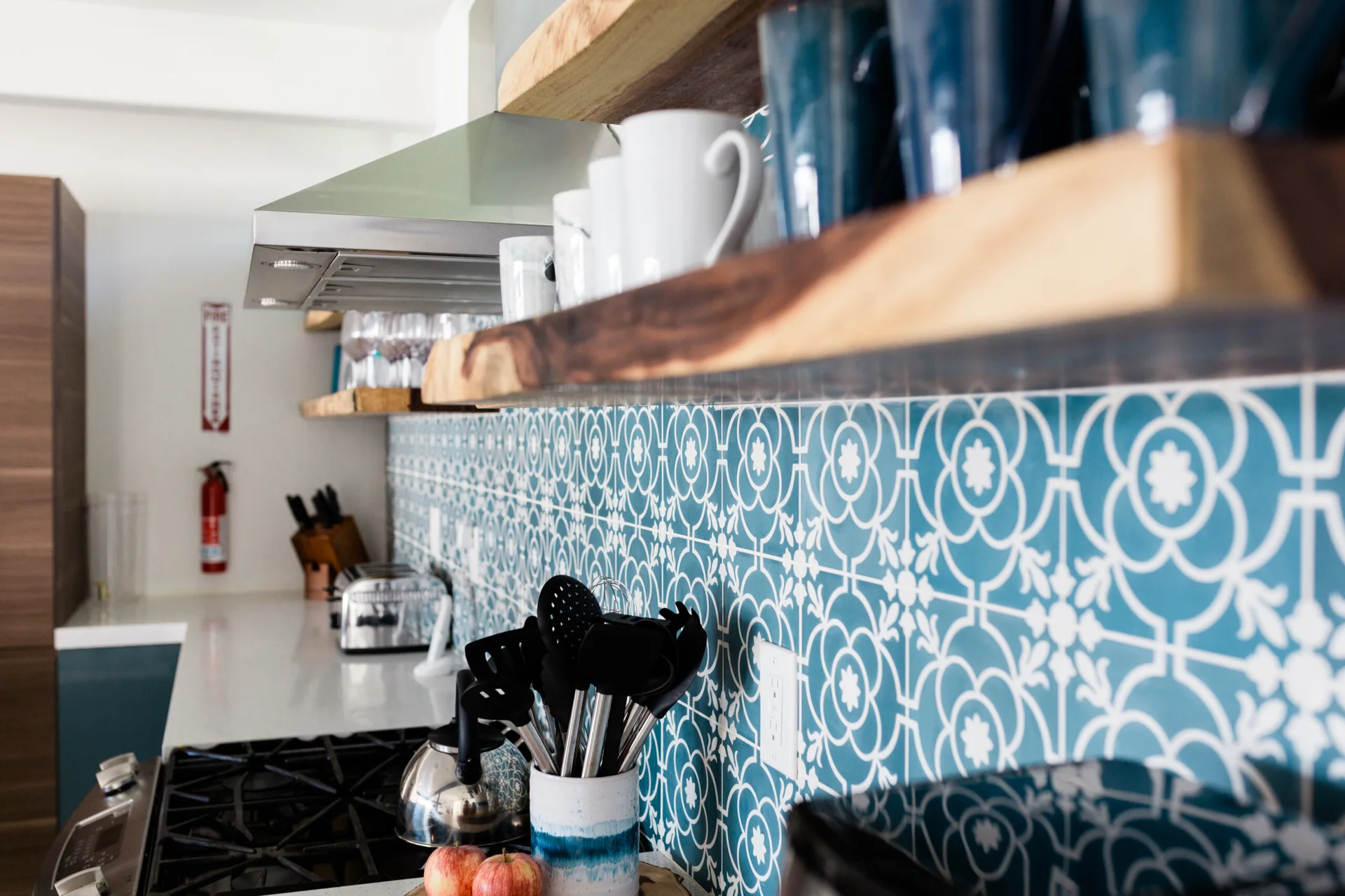 A kitchen counter view featuring a teal geometric tile backsplash, two live-edge natural wood open shelves, and kitchen utensils.