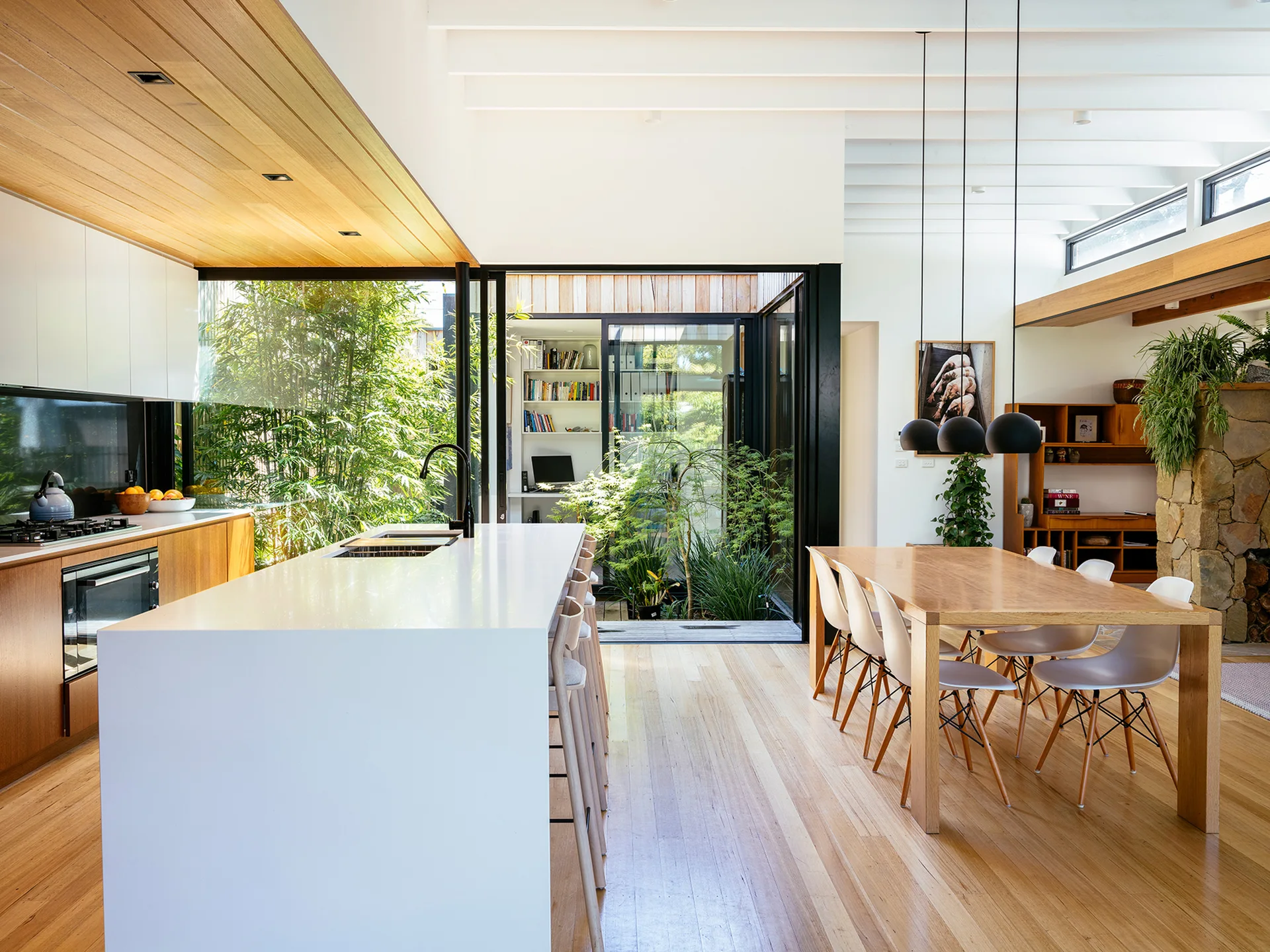 A modern open-plan kitchen and dining room features a white waterfall island, a wooden dining table under black pendant lights, and a central glass courtyard.