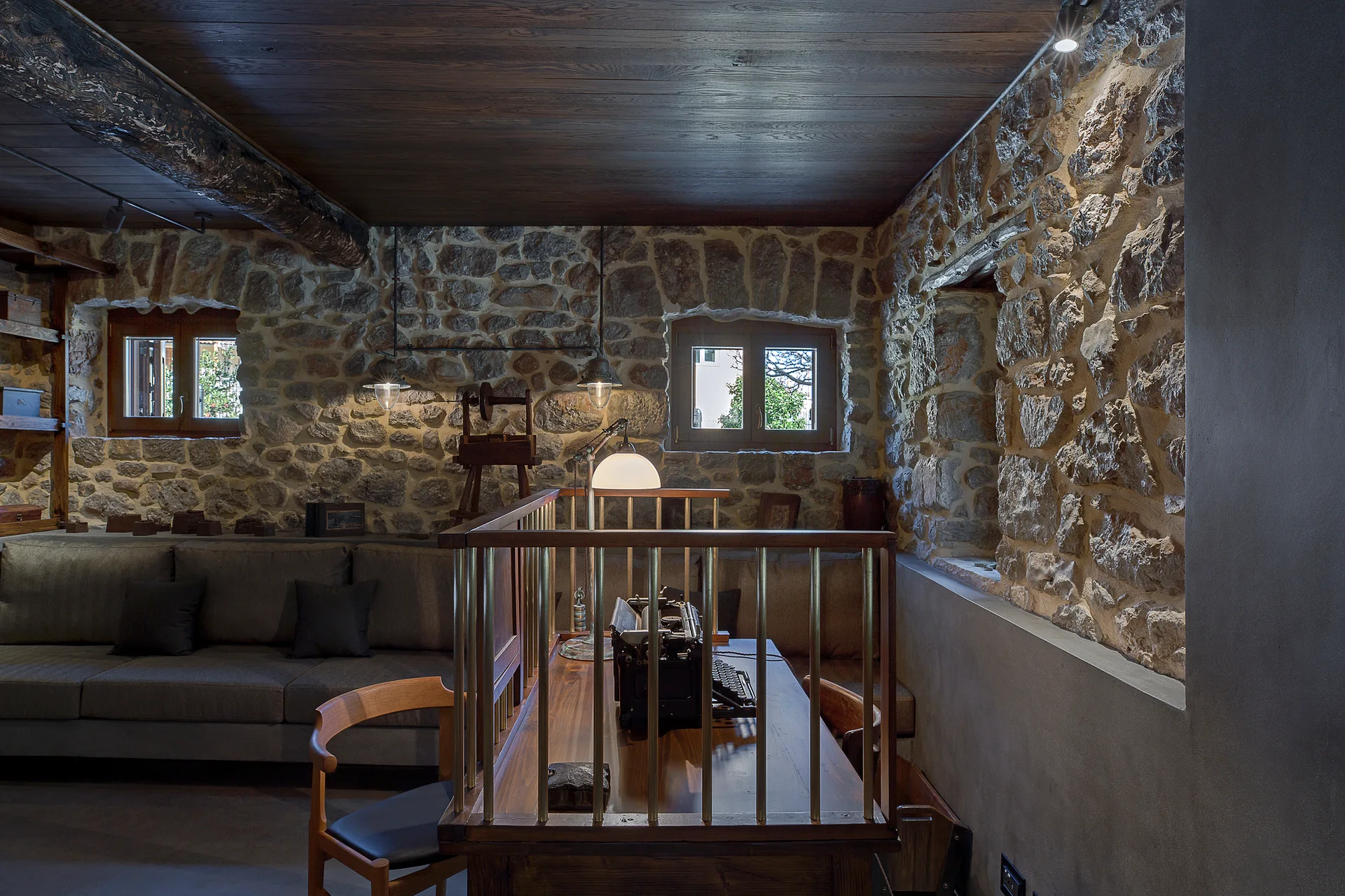 A rustic home office features a wooden desk with a typewriter, rough exposed stone walls, small windows, and a dark wood ceiling.