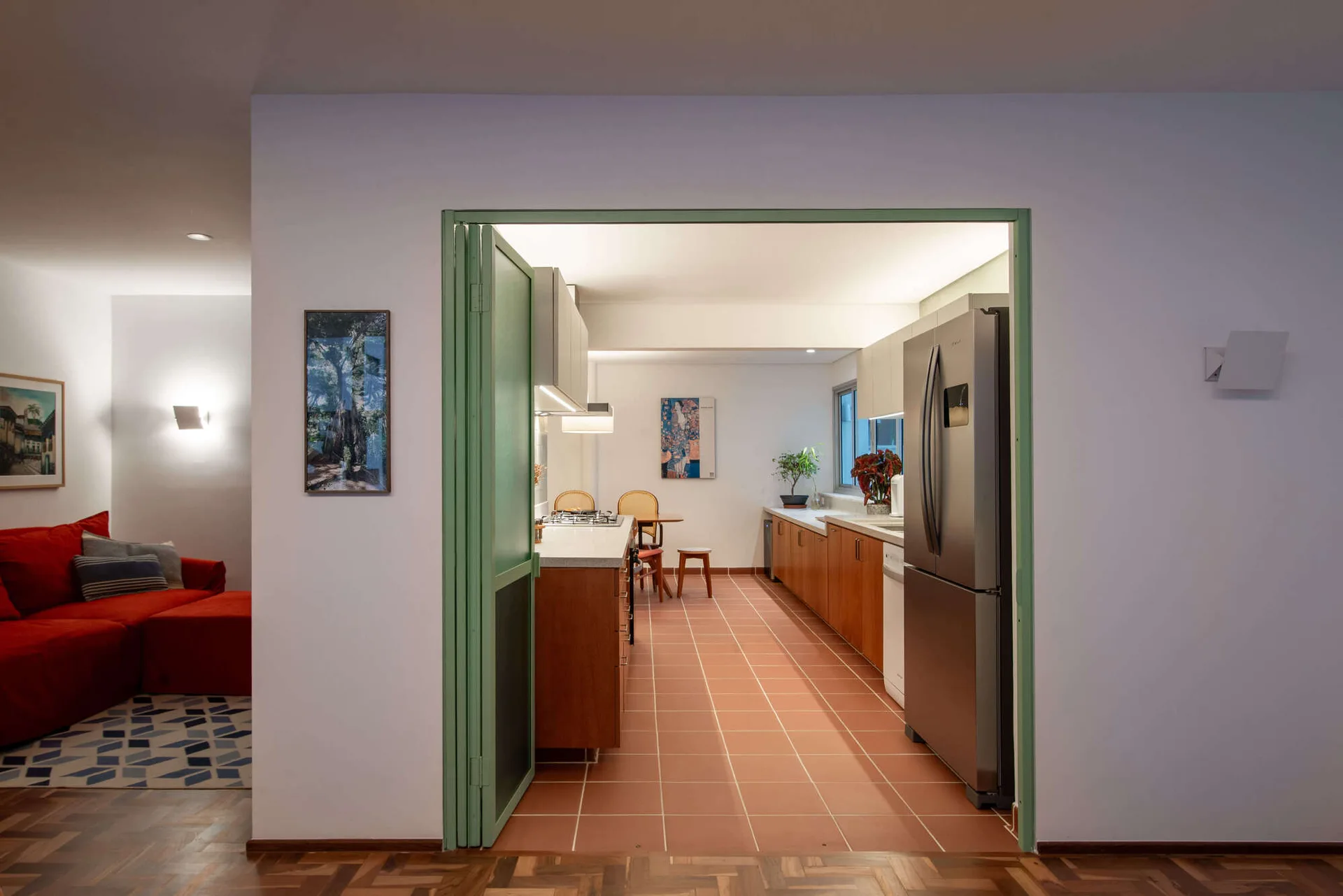 A view through a green-framed doorway into a galley kitchen featuring terracotta tile floors, wood base cabinets, and a stainless steel refrigerator.