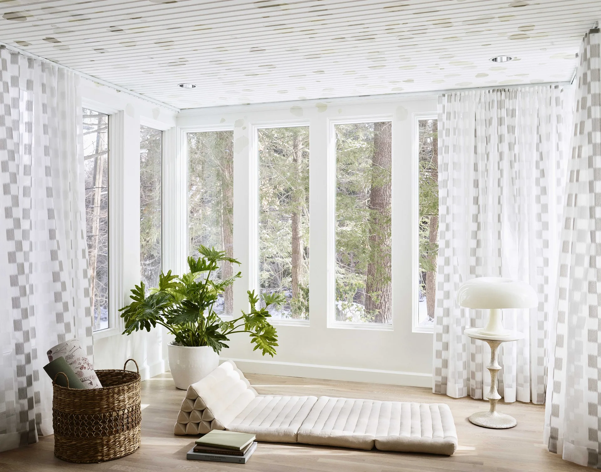 A bright wellness room features a low beige floor lounger, checkered curtains, and large windows looking out onto a snow-dusted forest.