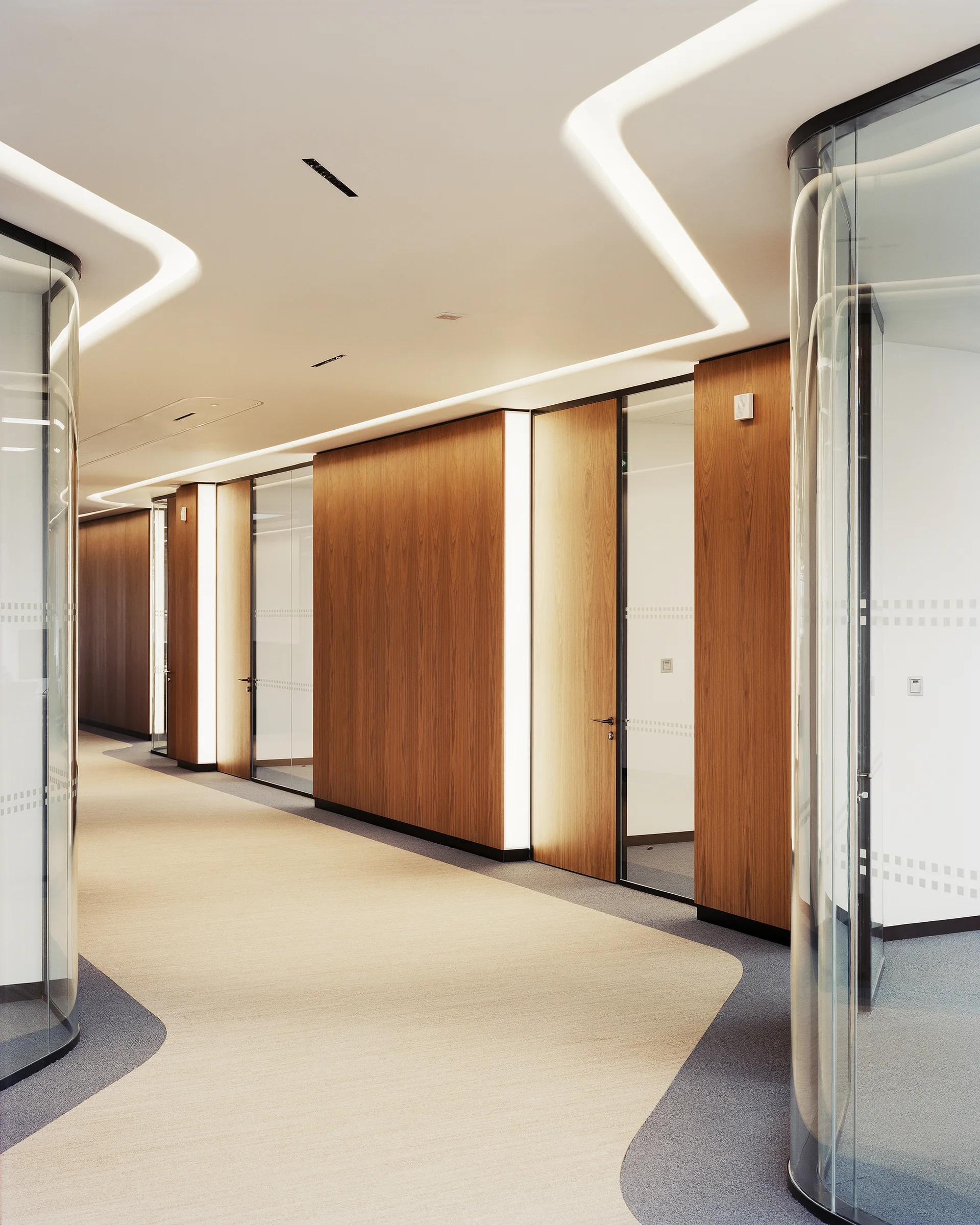 A contemporary office hallway featuring curved glass partition walls, wood-paneled doors, two-toned carpet, and recessed linear ceiling lights.