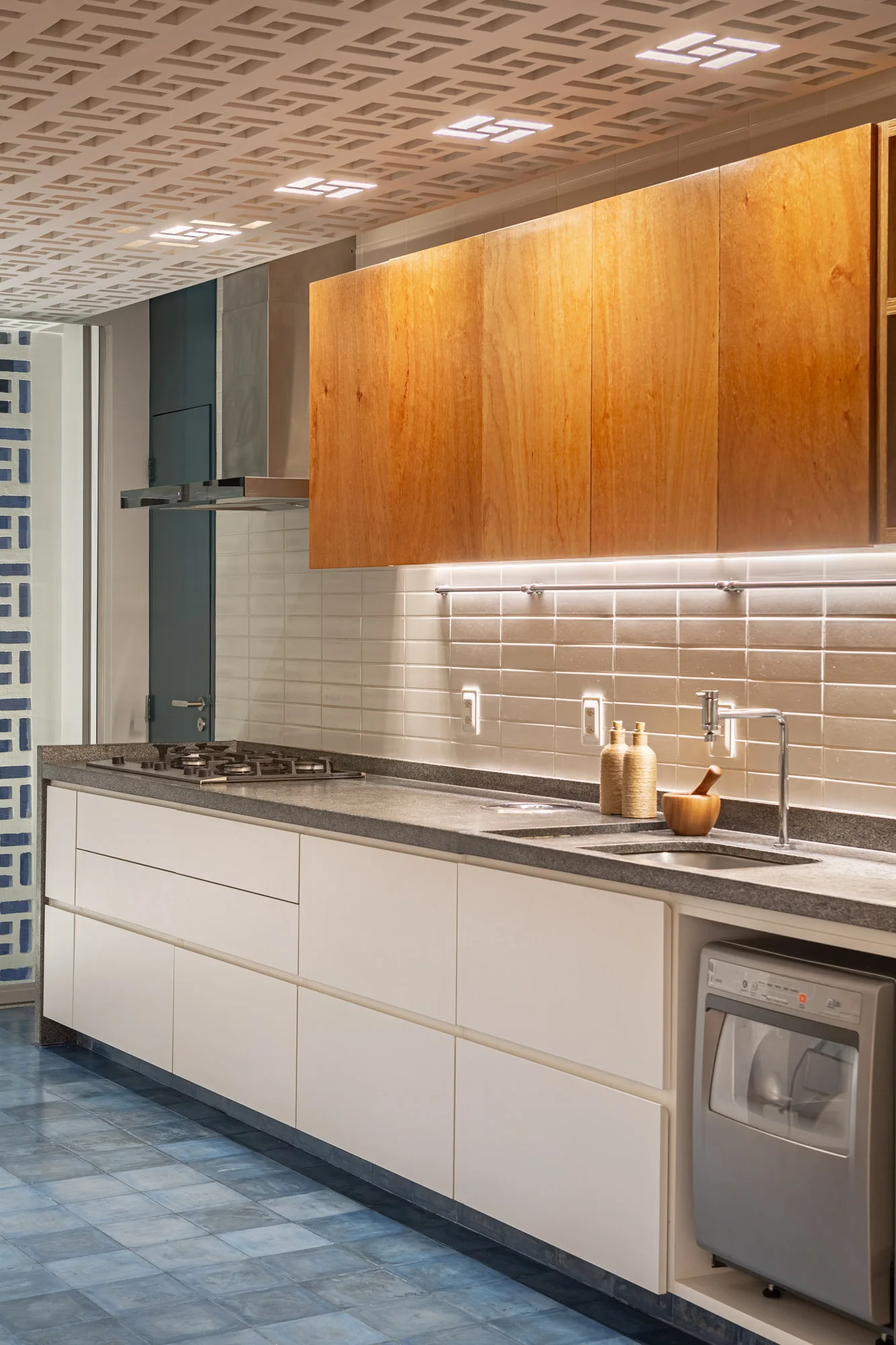 A modern kitchen with wood upper cabinets, white lower drawers, a grey stone countertop, and blue tile floors beneath a geometric patterned ceiling.
