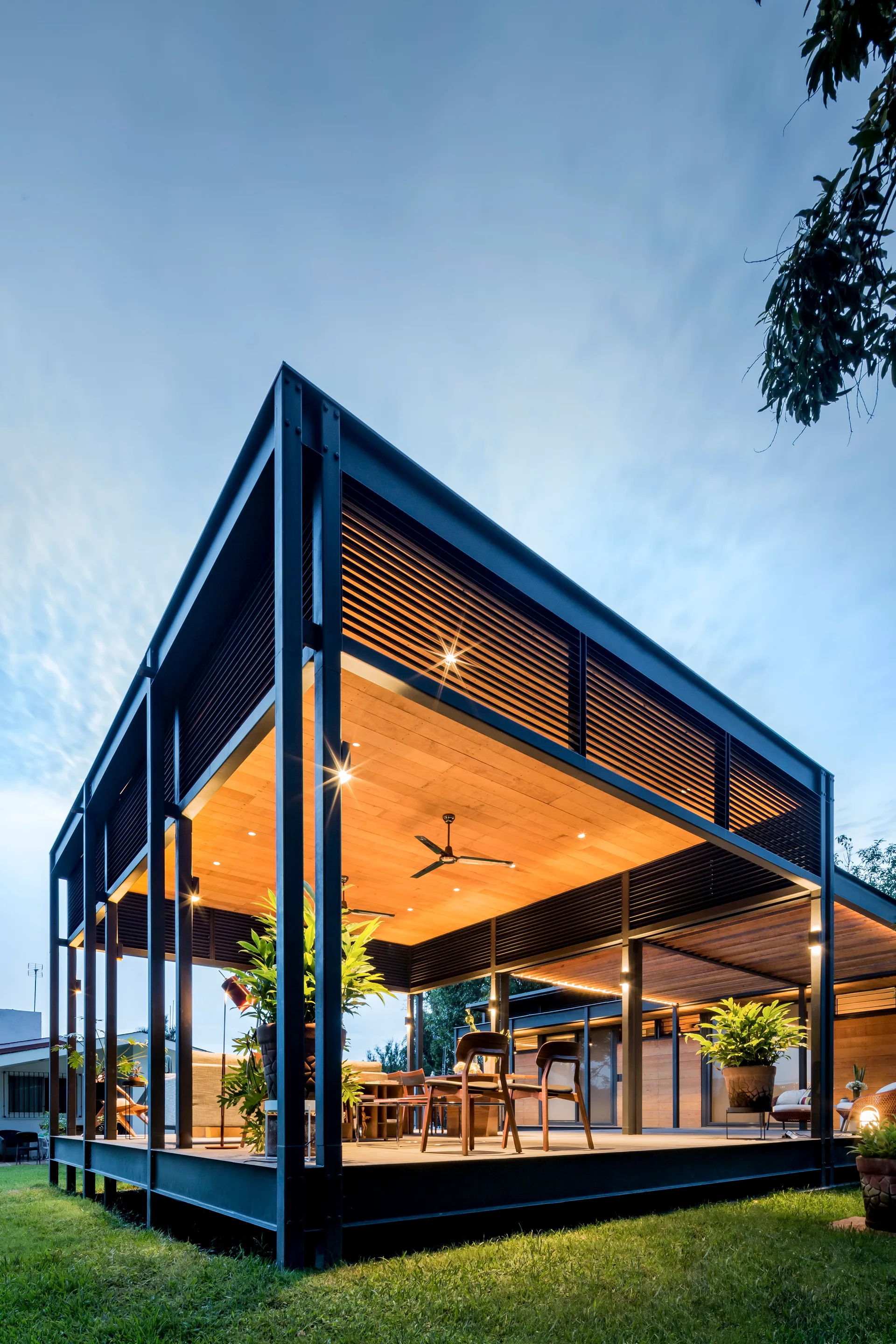 An elevated modern patio structure with a black steel frame, warm wood ceiling panels, and black slatted screens overlooking a grassy lawn.