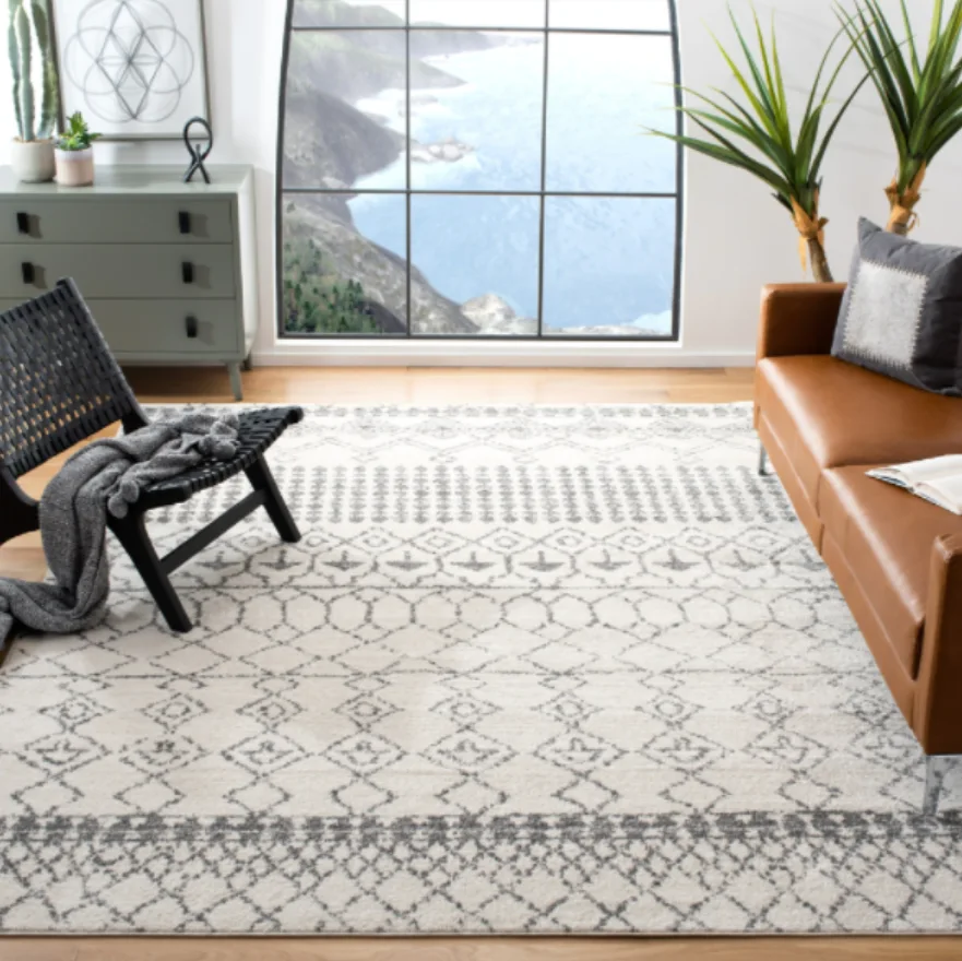 A modern living room features a tan leather sofa, a large cream and grey geometric rug, and a black woven accent chair near a large window.