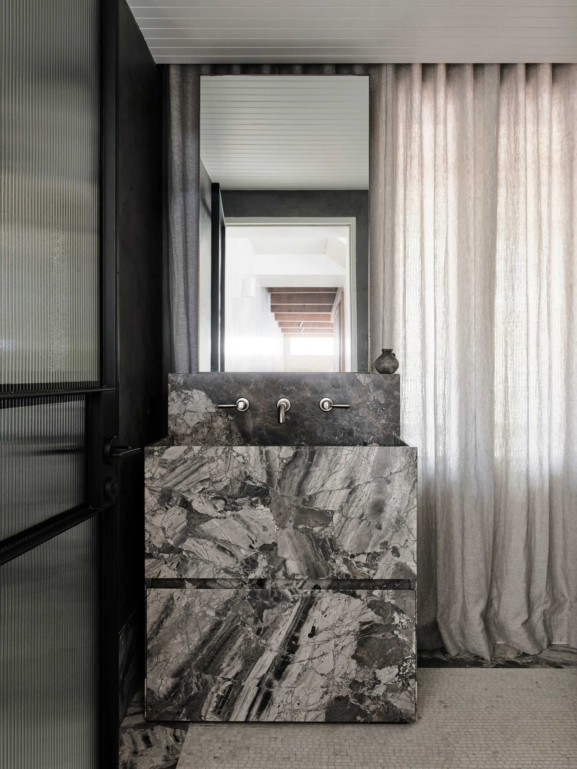 A modern bathroom features a grey veined marble vanity with a wall-mounted faucet, set against dark walls and a sheer beige curtain.