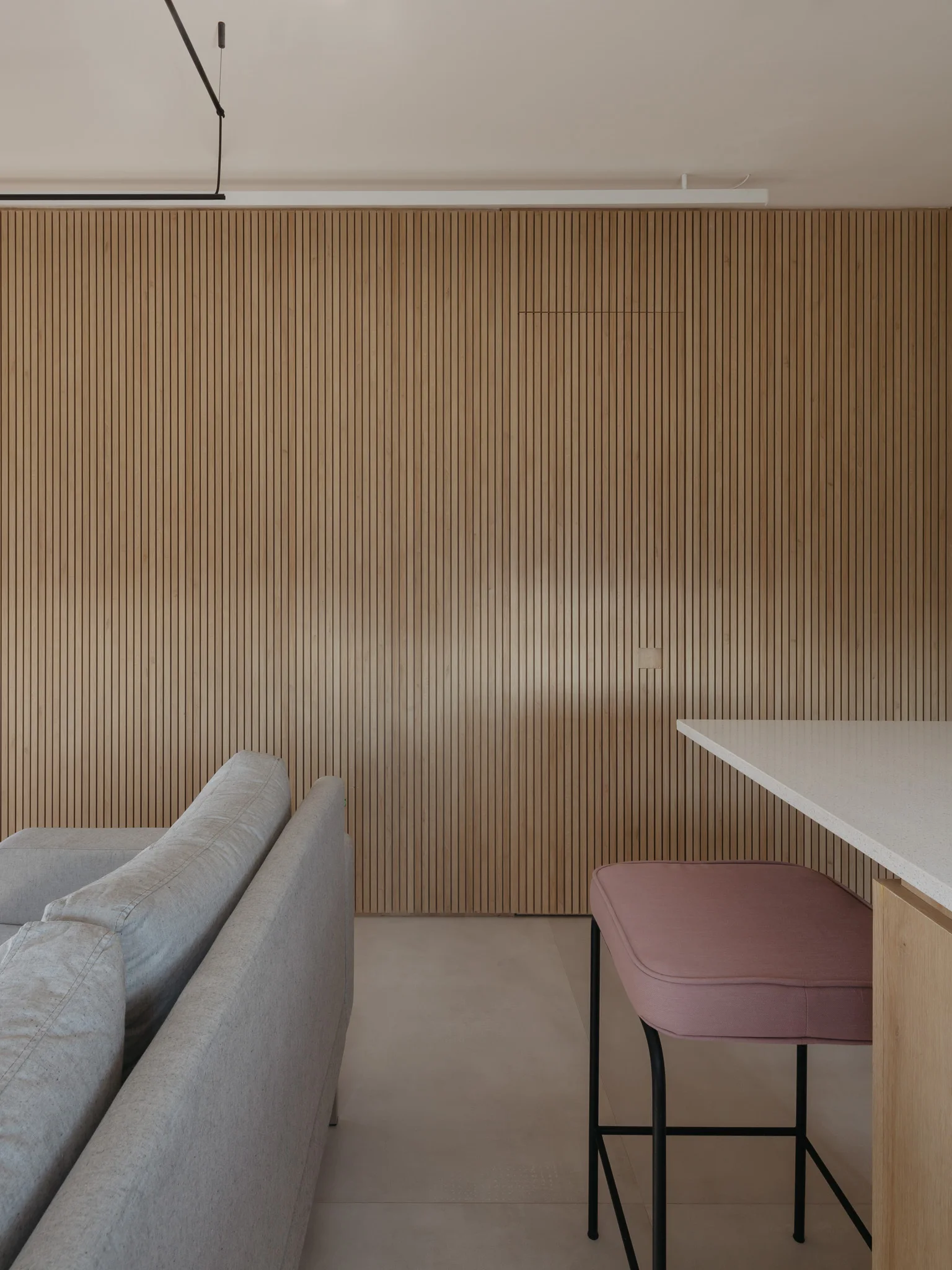 A gray fabric sofa and a pink upholstered bar stool at a white counter in front of a floor-to-ceiling vertical slatted wood accent wall.