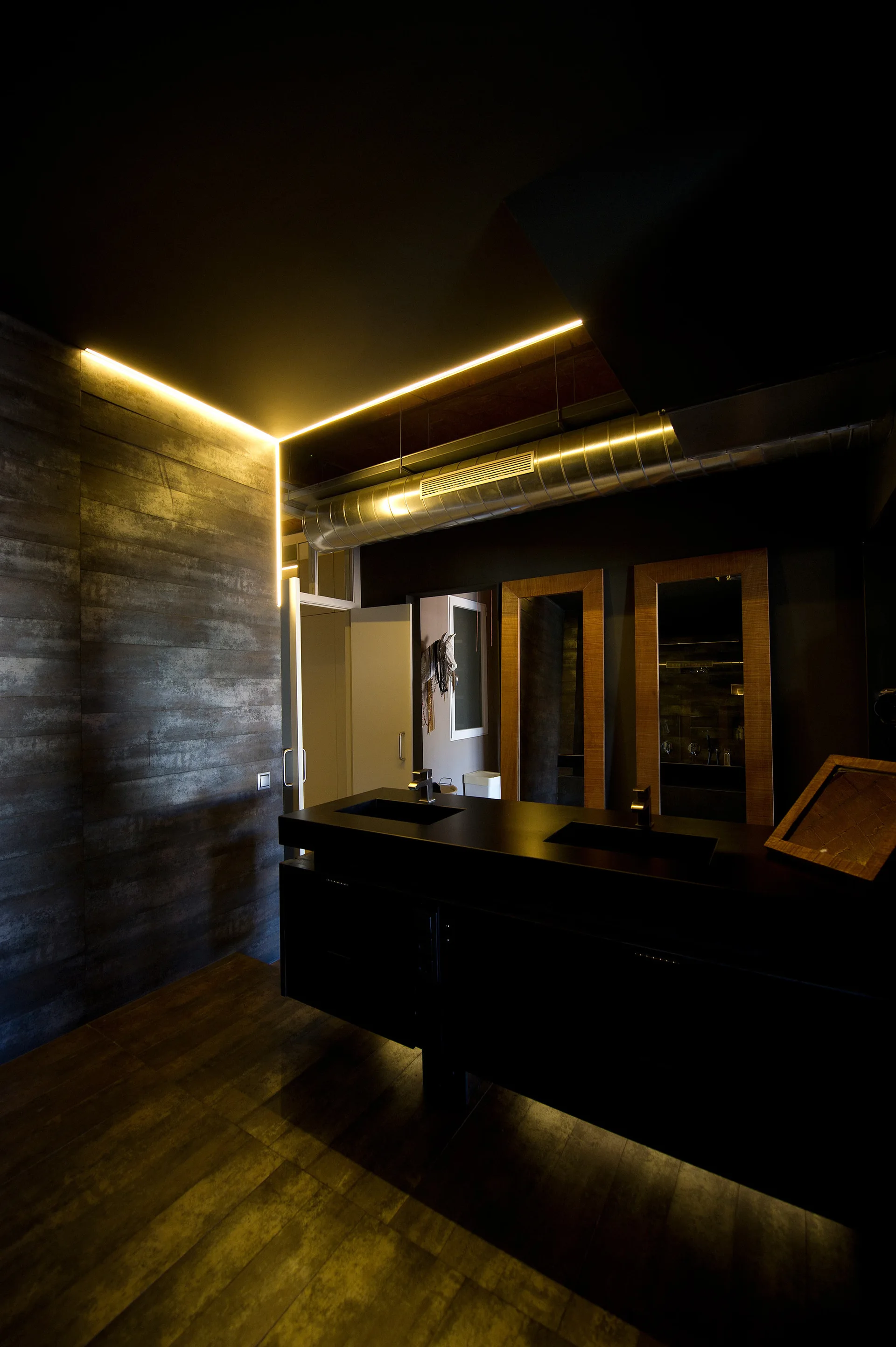 A dark industrial bathroom features a matte black double vanity, two wood-framed mirrors, and bright yellow cove lighting along a textured wall.