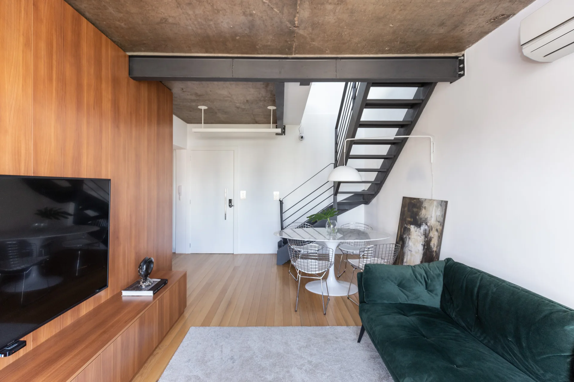 A modern open-plan living room features a green tufted sofa, a wood-paneled wall, and a black steel staircase beneath an exposed concrete ceiling.