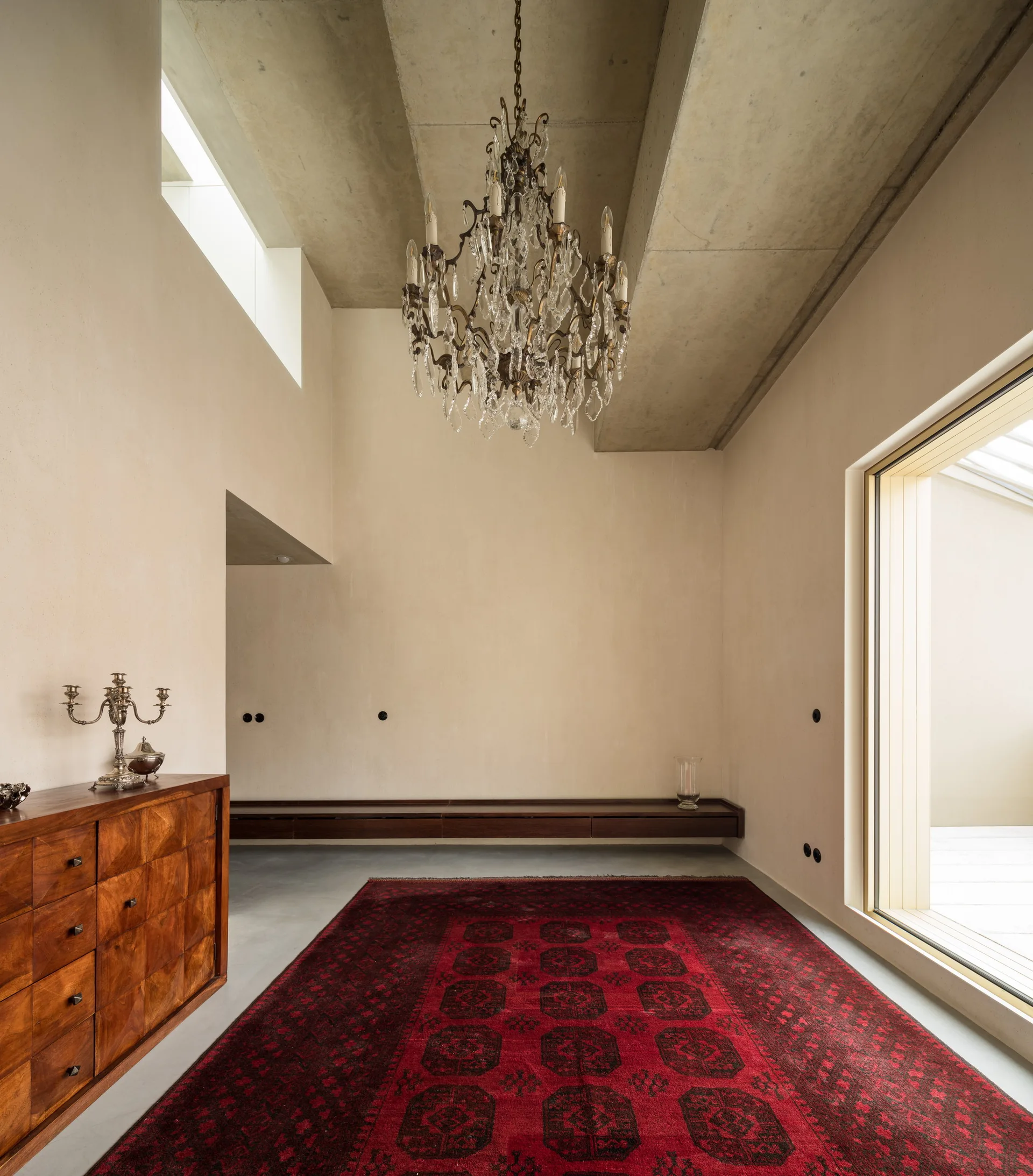 A minimalist room features a raw concrete ceiling with a crystal chandelier, cream walls, and a large red patterned rug on a grey floor.