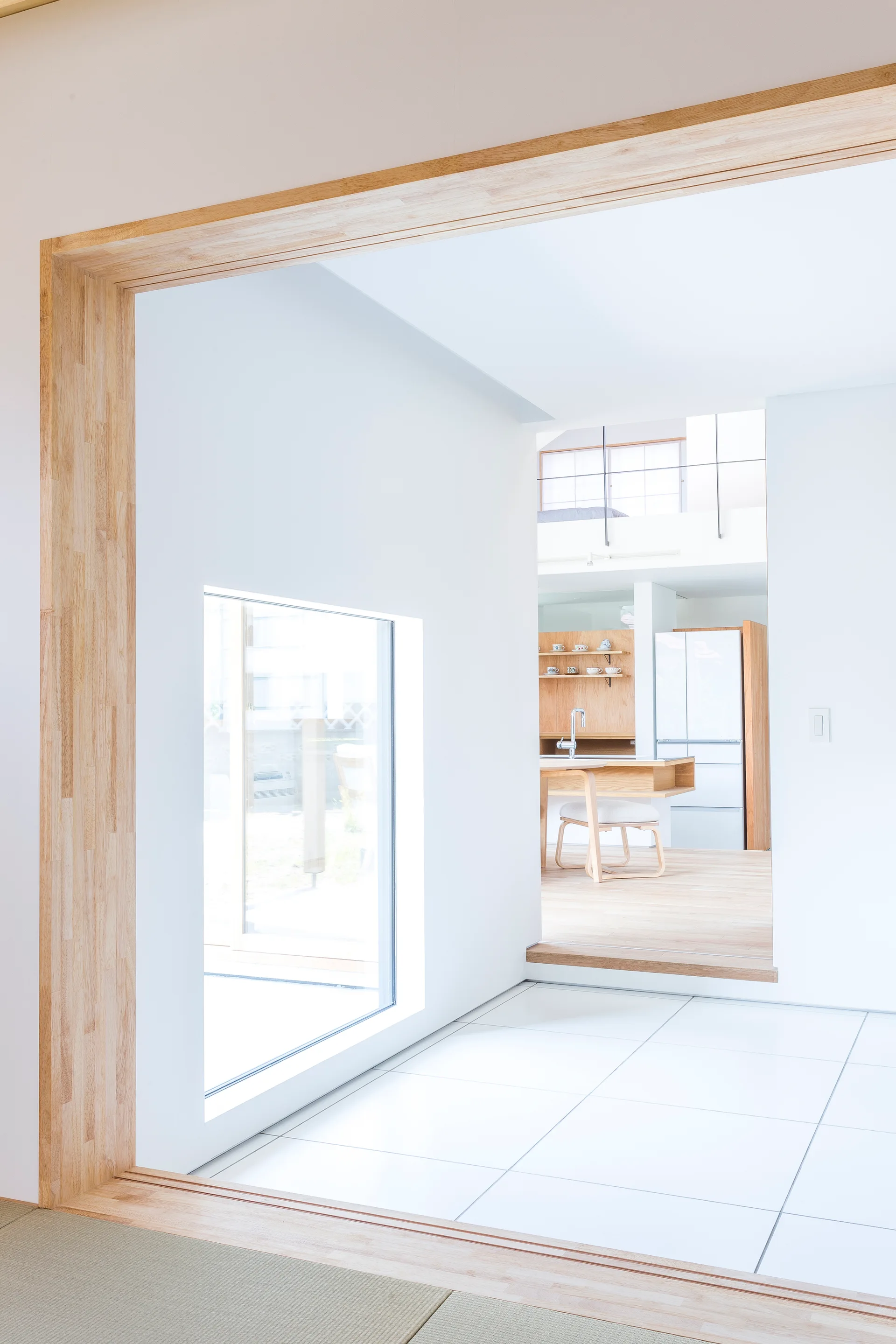 An open interior view features a light wood doorframe leading past a tiled floor with a square window toward a kitchen with wood floors.