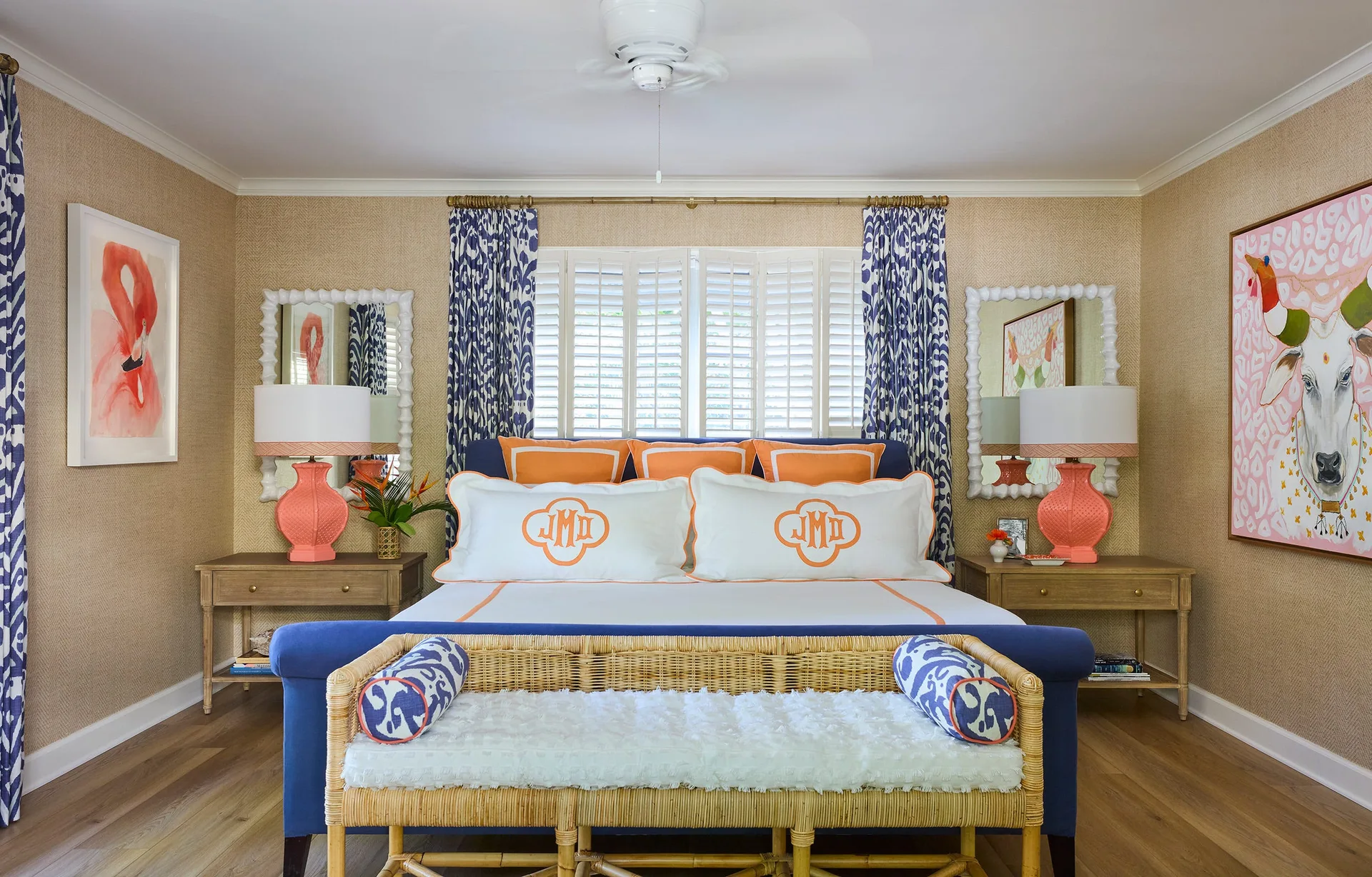 A symmetrical bedroom features a blue upholstered bed flanked by wooden nightstands with coral lamps, white mirrors, and a rattan bench at the foot.