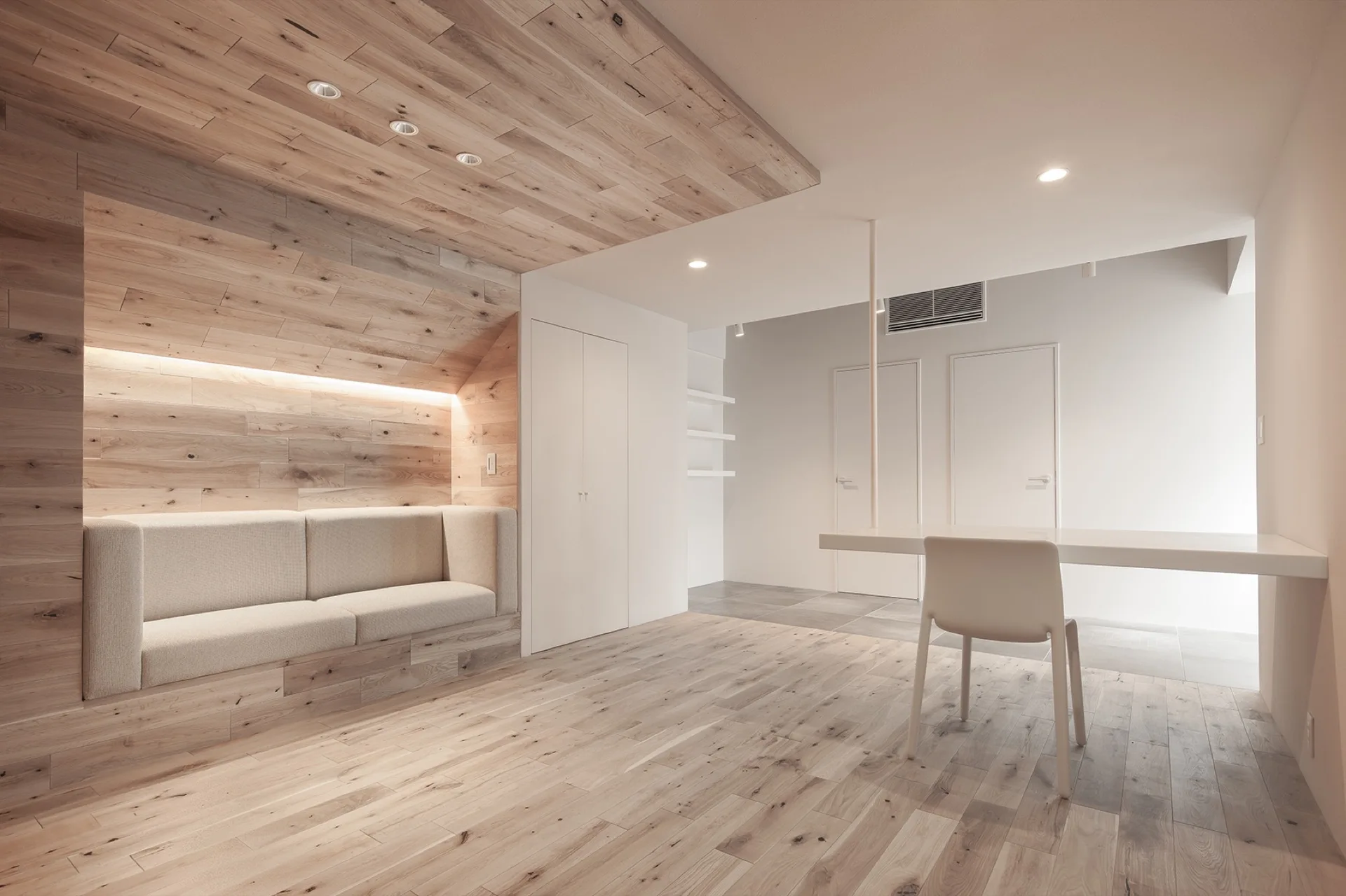 A minimalist room features a built-in beige sofa nested inside a light wood-paneled alcove opposite a wall-mounted white table with a single chair.