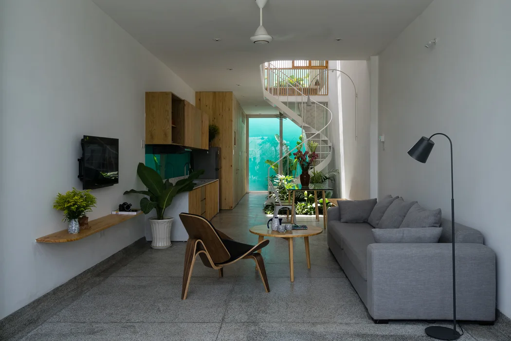 A narrow minimalist living room features a gray sofa, molded wood chair, and a white spiral staircase leading upward in the background.