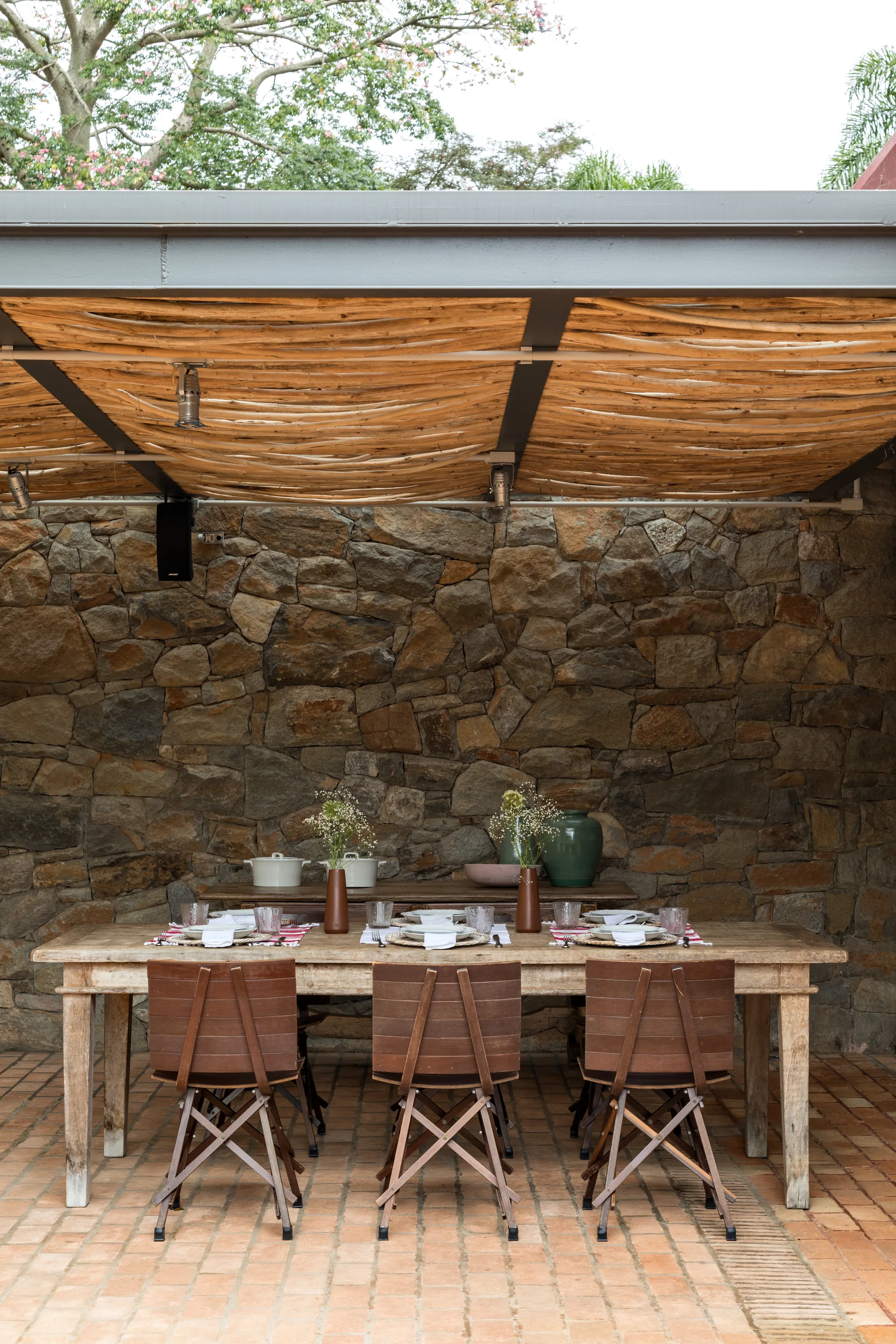 A rustic outdoor dining table and chairs sit on a brick patio under a textured branch roof against a rugged stone wall.